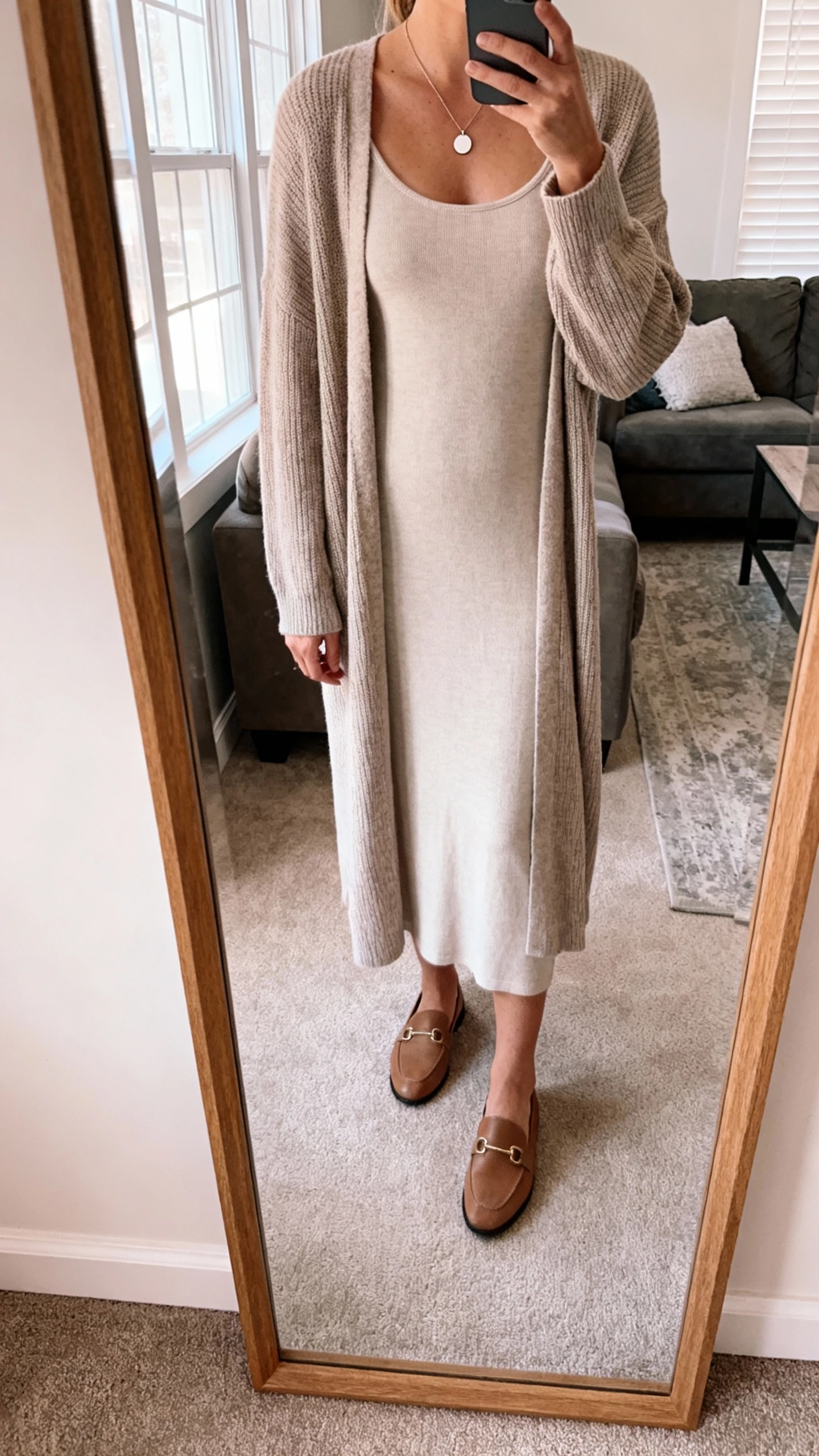 iPhone mirror selfie of a woman wearing a column dress with a longline cardigan, minimalist pendant necklace, and loafers, face not visible, cropped at shoulders, living room mirror, gentle window light, iPhone photo quality