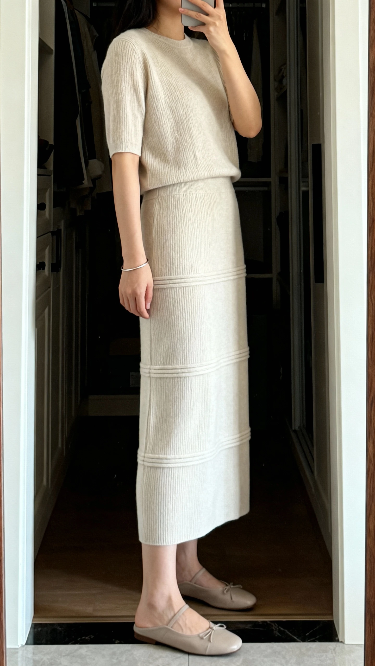 iPhone mirror selfie of a woman wearing a column knit skirt set with modest layers and sleek lines (matching knit top and straight knit skirt), understated flats and a thin bracelet, face not visible, side angle, closet full-length mirror, gentle indoor light, iPhone photo quality