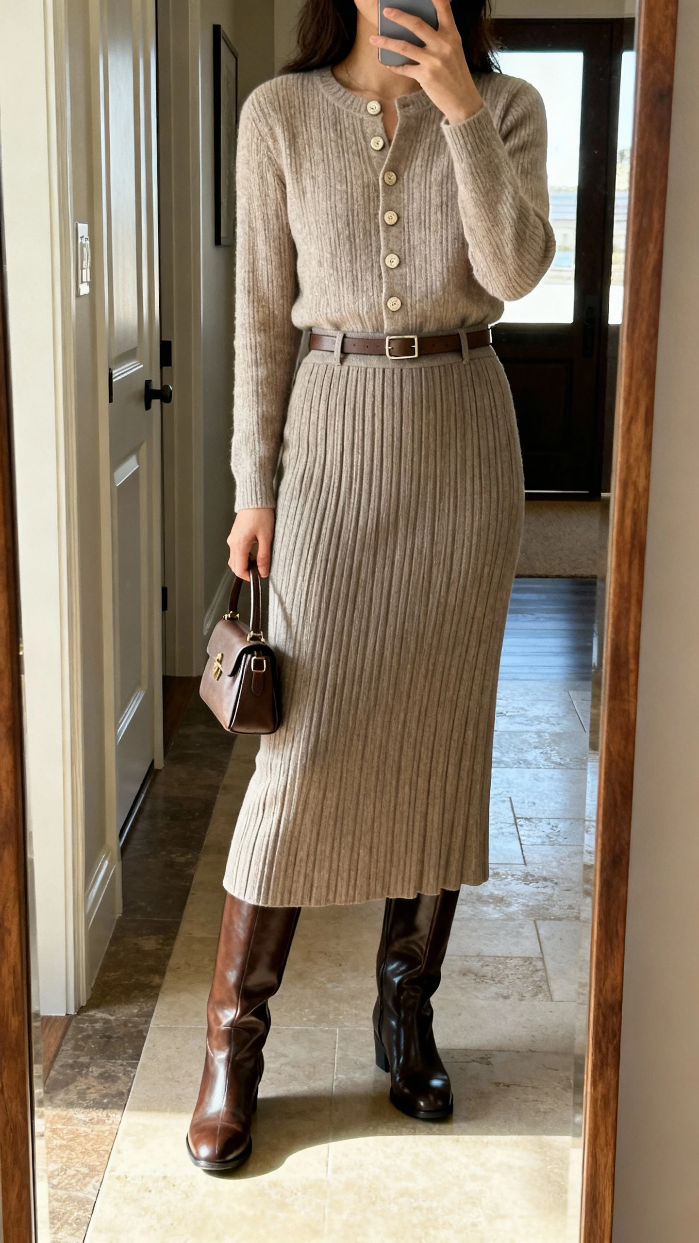 iPhone mirror selfie of a woman wearing a column skirt with a buttoned knit top and polished tall boots, minimal belt and small top-handle bag, face not visible, cropped at neck, entryway full-length mirror, natural daylight, iPhone photo quality.