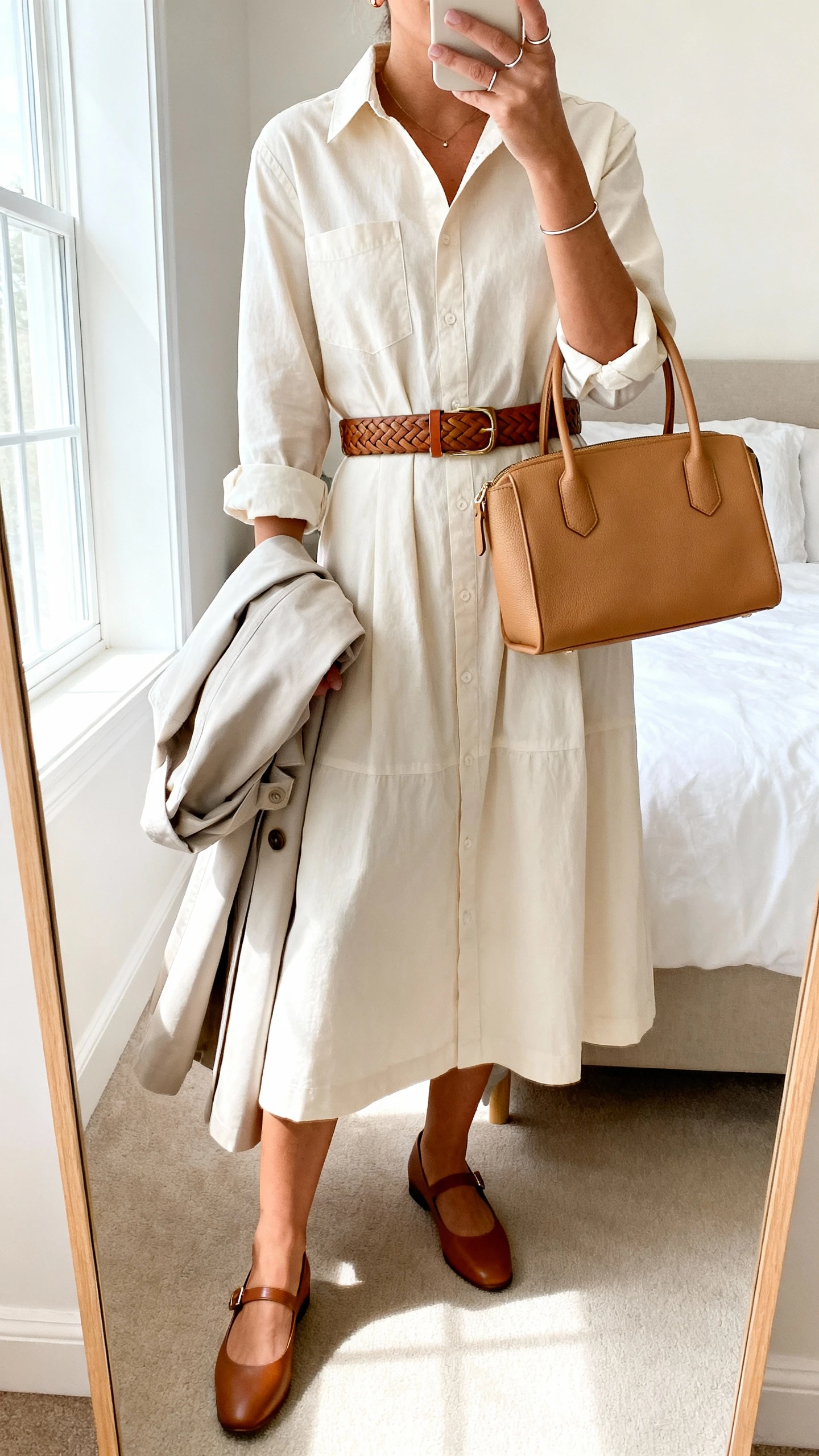 iPhone mirror selfie of a woman wearing a cotton poplin shirt dress with a midi hem and cuffed sleeves, a woven leather belt, refined Mary Janes, simple studs with a slim ring, and a top-handle satchel in tan, optional light trench coat folded over her arm, face not visible, cropped at shoulders, bedroom mirror, bright window light, iPhone photo quality.