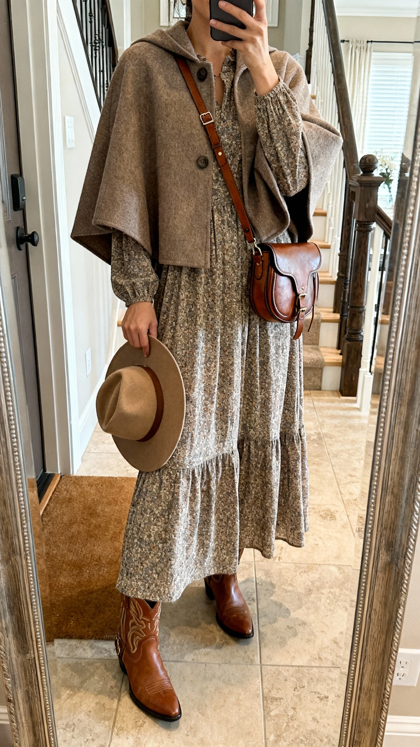 iPhone mirror selfie of a woman wearing a country-chic maxi dress with a cape-coat, western-style leather boots, a felt hat in hand, and a crossbody saddle bag, face not visible, cropped at neck, entryway mirror, natural indoor daylight, iPhone photo quality.