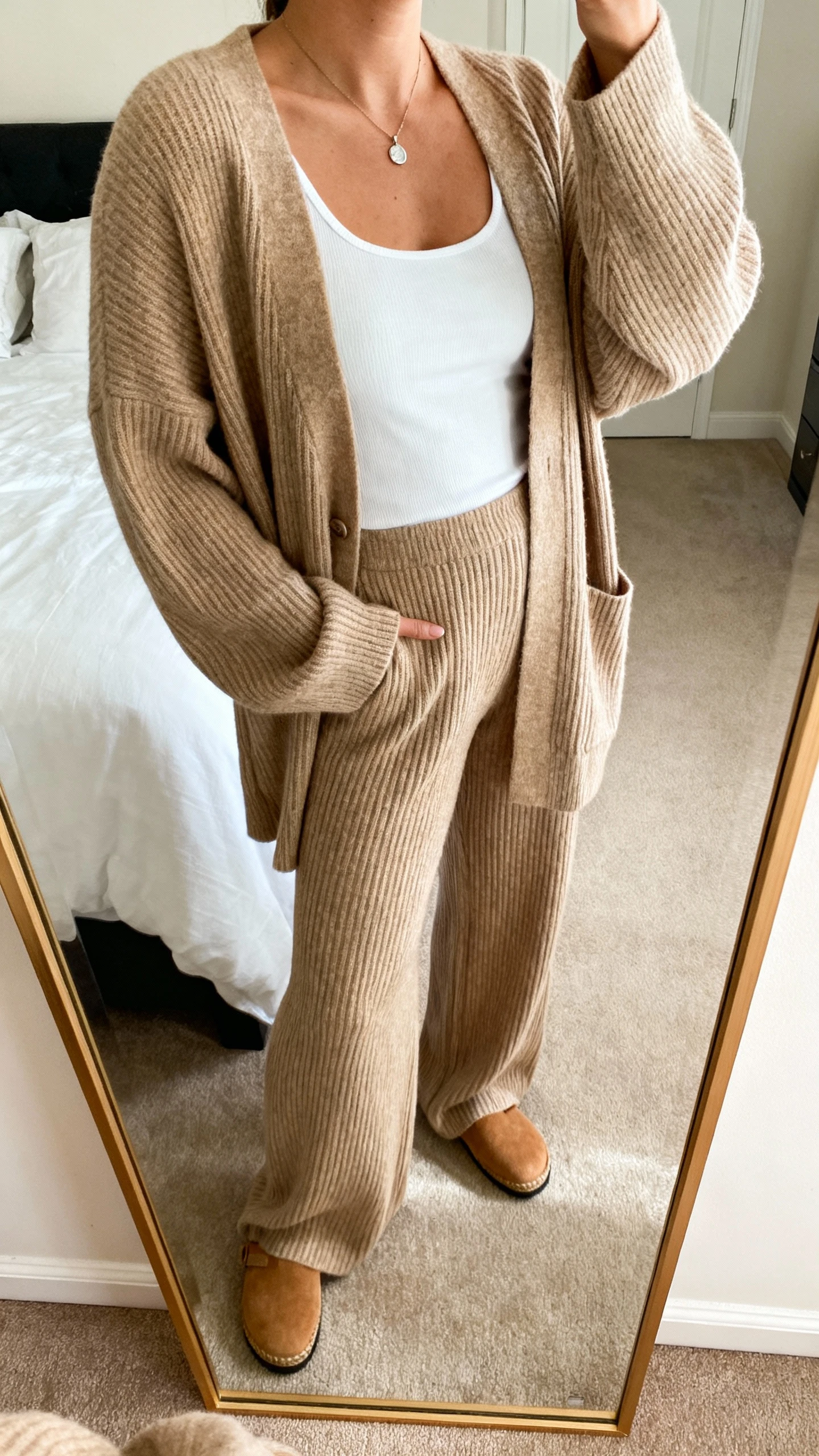 iPhone mirror selfie of a woman wearing a cozy ribbed knit set: relaxed cardigan and matching wide-leg knit pants in sand, with a white tank underneath, tan slip-on mules, and a simple pendant necklace, face not visible, cropped at neck, bedroom mirror, warm indoor lighting, iPhone photo quality.