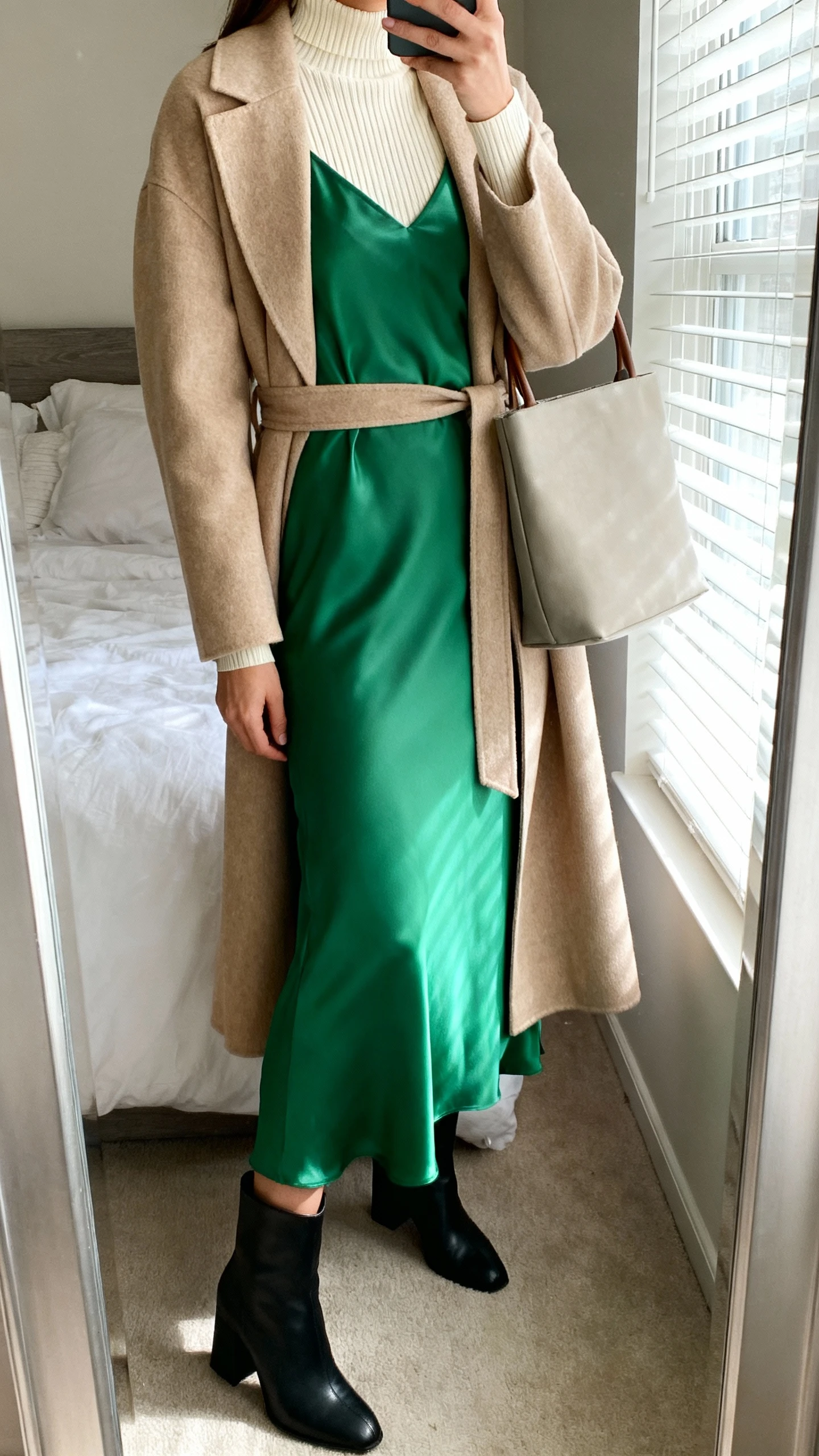 iPhone mirror selfie of a woman wearing a cream fine-gauge mock-neck under a satin-soft emerald maxi dress with modest neckline, topped with a beige belted duster coat, black block-heel boots, and a simple tote, face not visible, cropped at neck, bedroom mirror, morning window light, iPhone photo quality.
