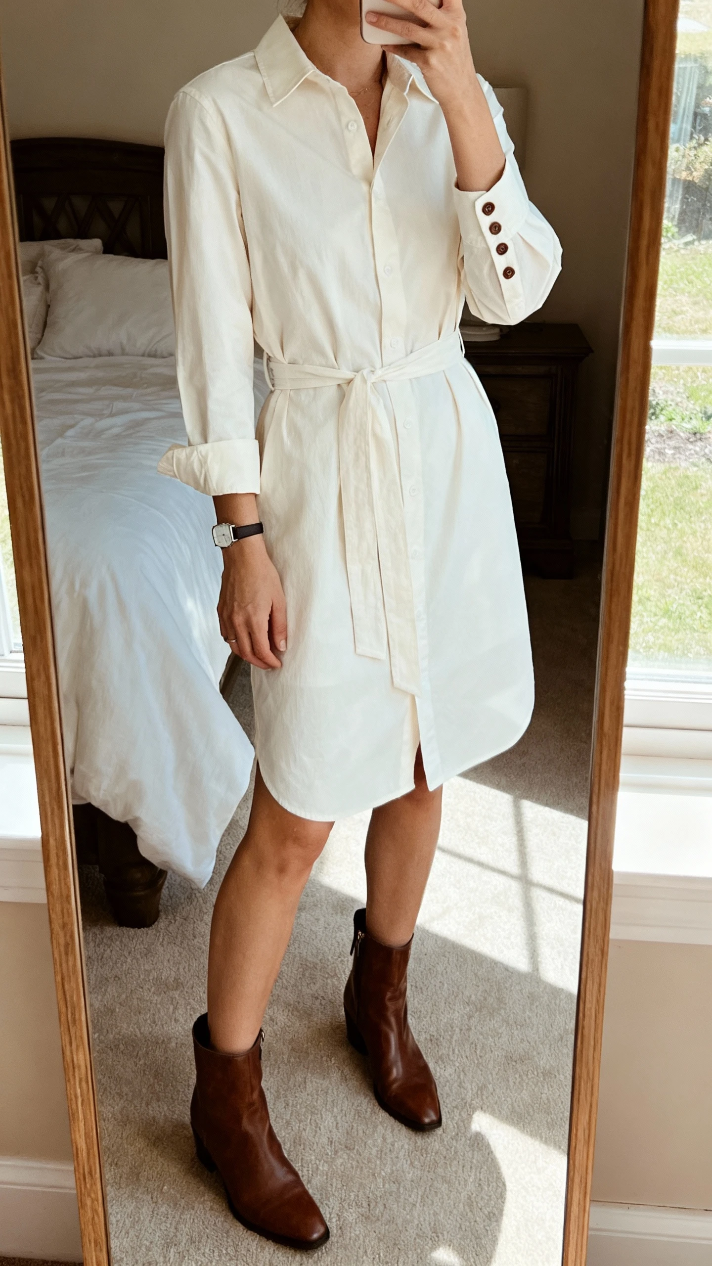 iPhone mirror selfie of a woman wearing a crisp shirt dress with a belted waist, long sleeves buttoned at the cuff, paired with leather ankle boots and a slim watch, face not visible, cropped at neck, bedroom mirror, soft afternoon window light, iPhone photo quality.