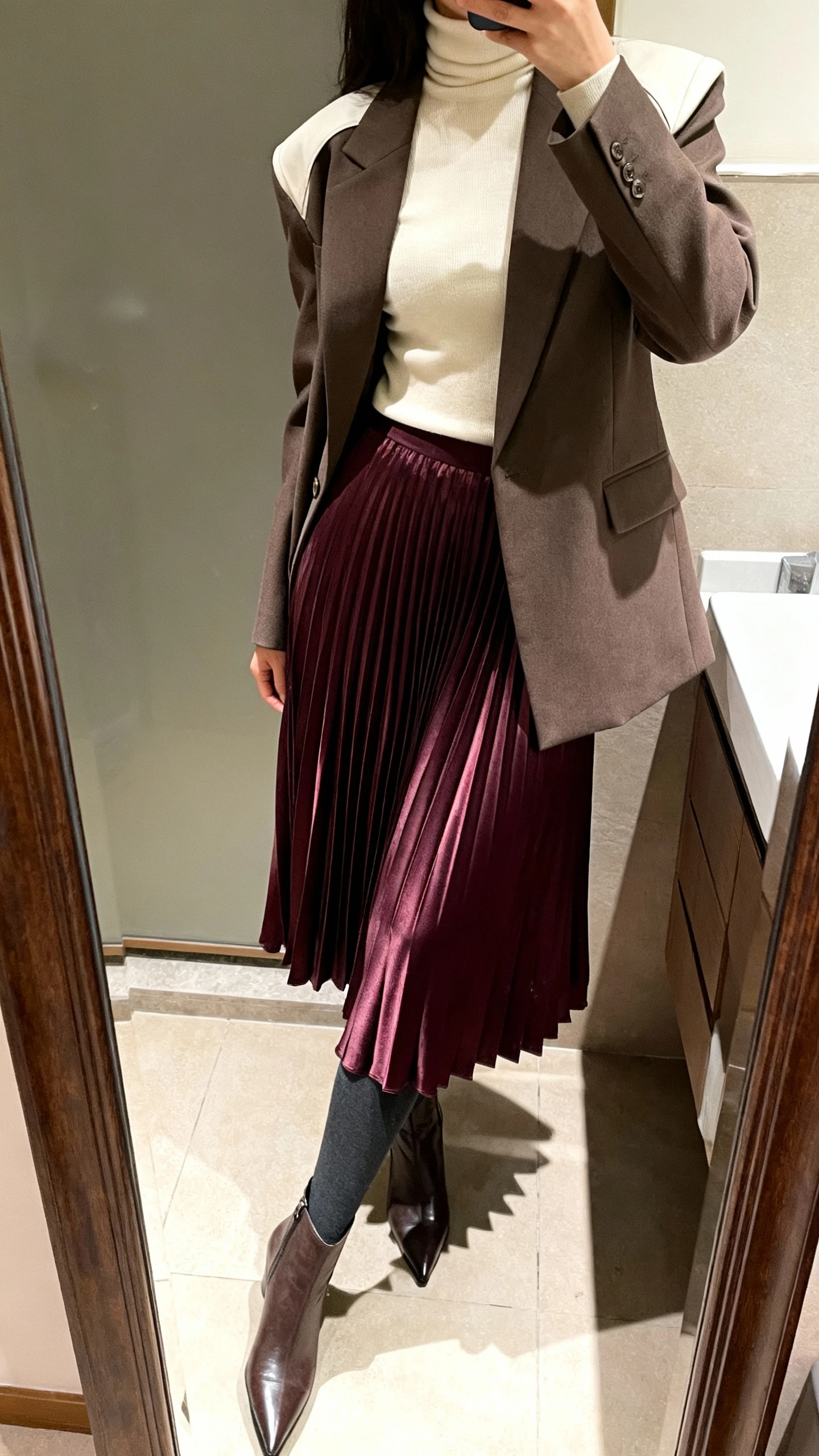 iPhone mirror selfie of a woman wearing a deep plum satin pleated midi skirt, cream fitted turtleneck, single-breasted blazer with light shoulder structure, opaque charcoal tights, and pointed-toe ankle boots, face not visible, side angle in a bathroom mirror, indoor lighting, iPhone photo quality.