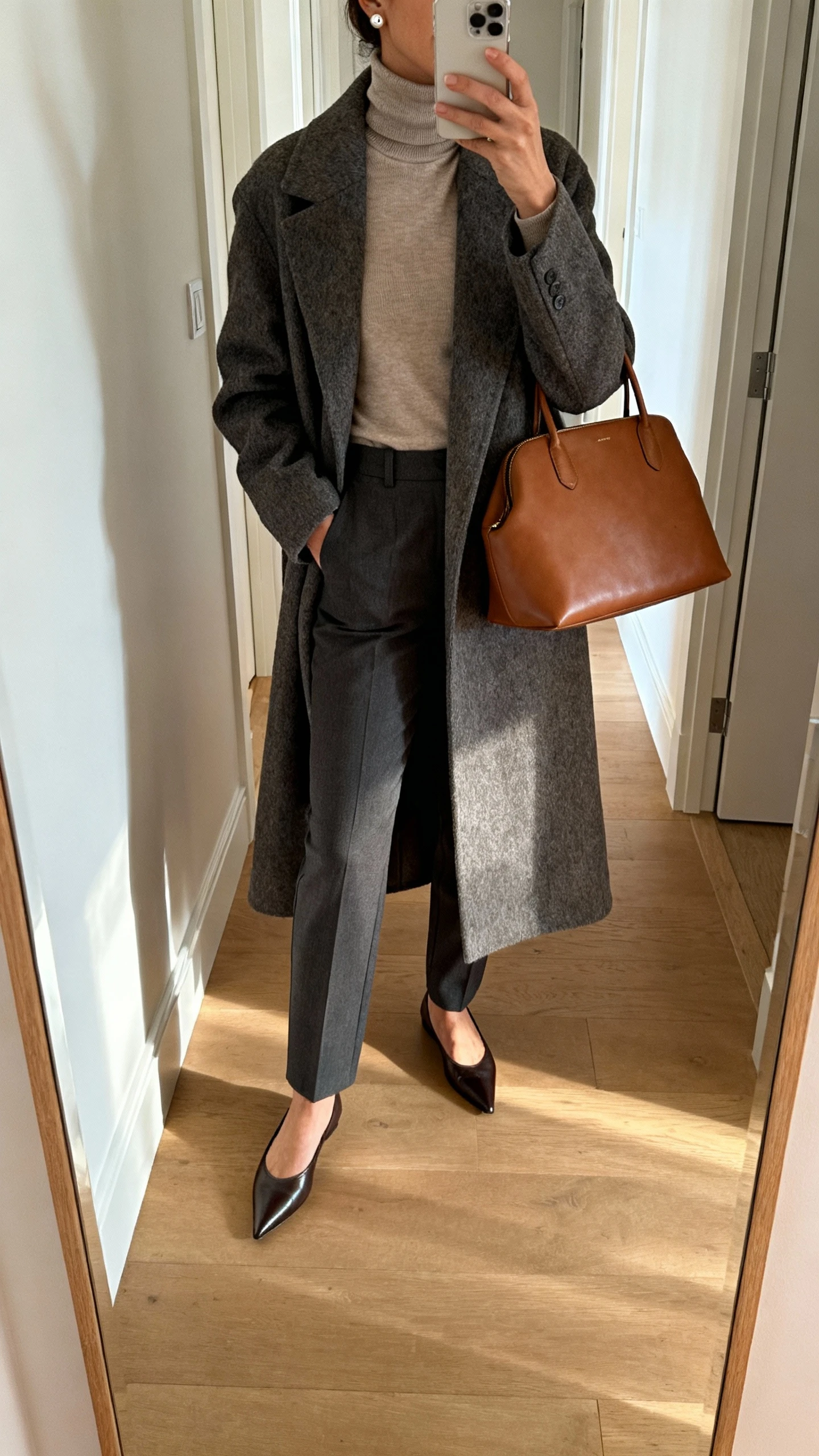iPhone mirror selfie of a woman wearing a fitted turtleneck under a long wool coat with tailored trousers, pointed flats, leather tote, and simple stud earrings, face not visible, cropped at shoulders, hallway mirror, soft morning light, iPhone photo quality