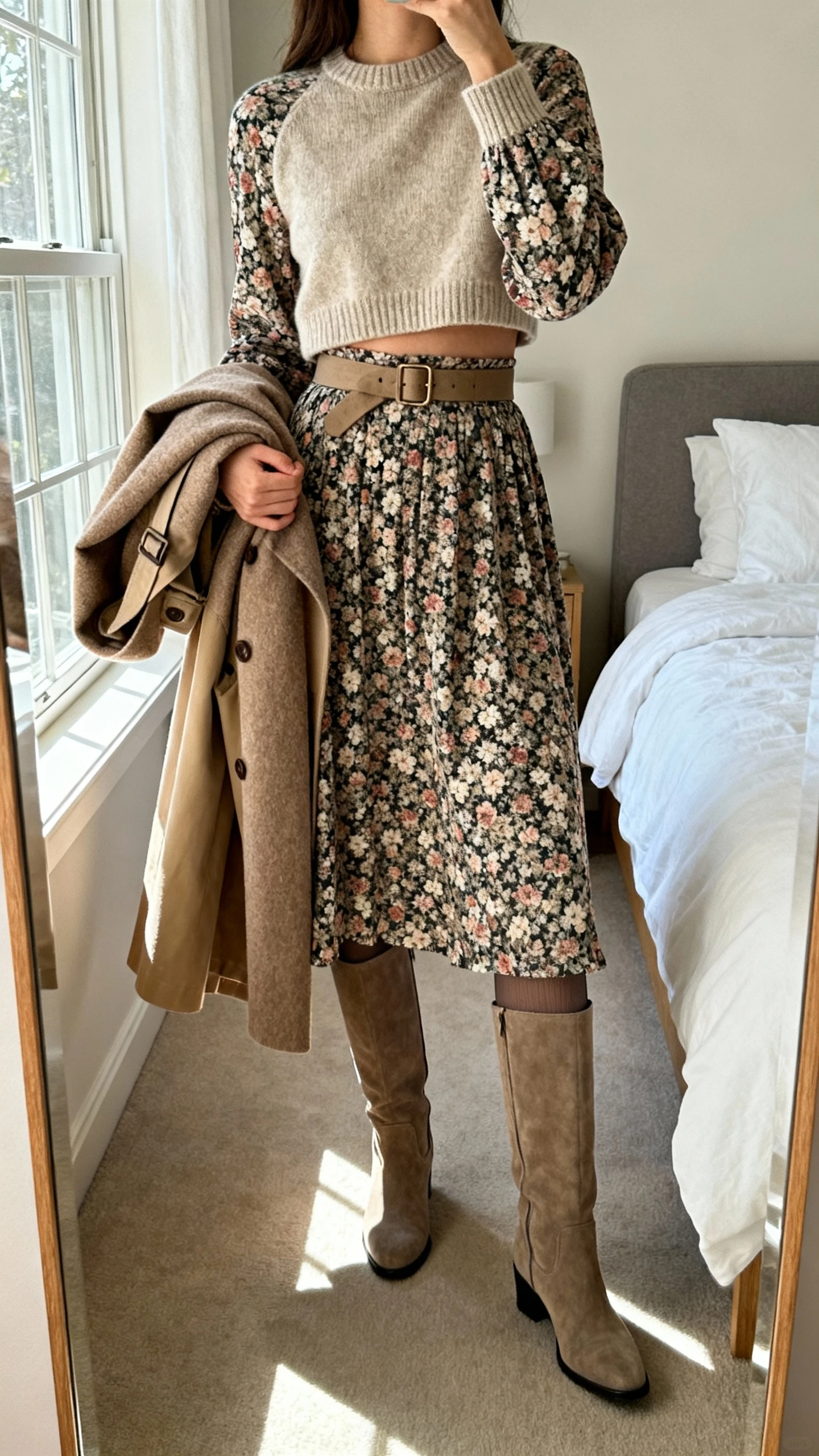 iPhone mirror selfie of a woman wearing a floral long-sleeve midi dress layered with a cropped crewneck sweater belted at the waist, opaque tights, tall boots, and a trench-style wool coat draped over the arm, face not visible, cropped at shoulders, bedroom mirror with natural window light, iPhone photo quality.
