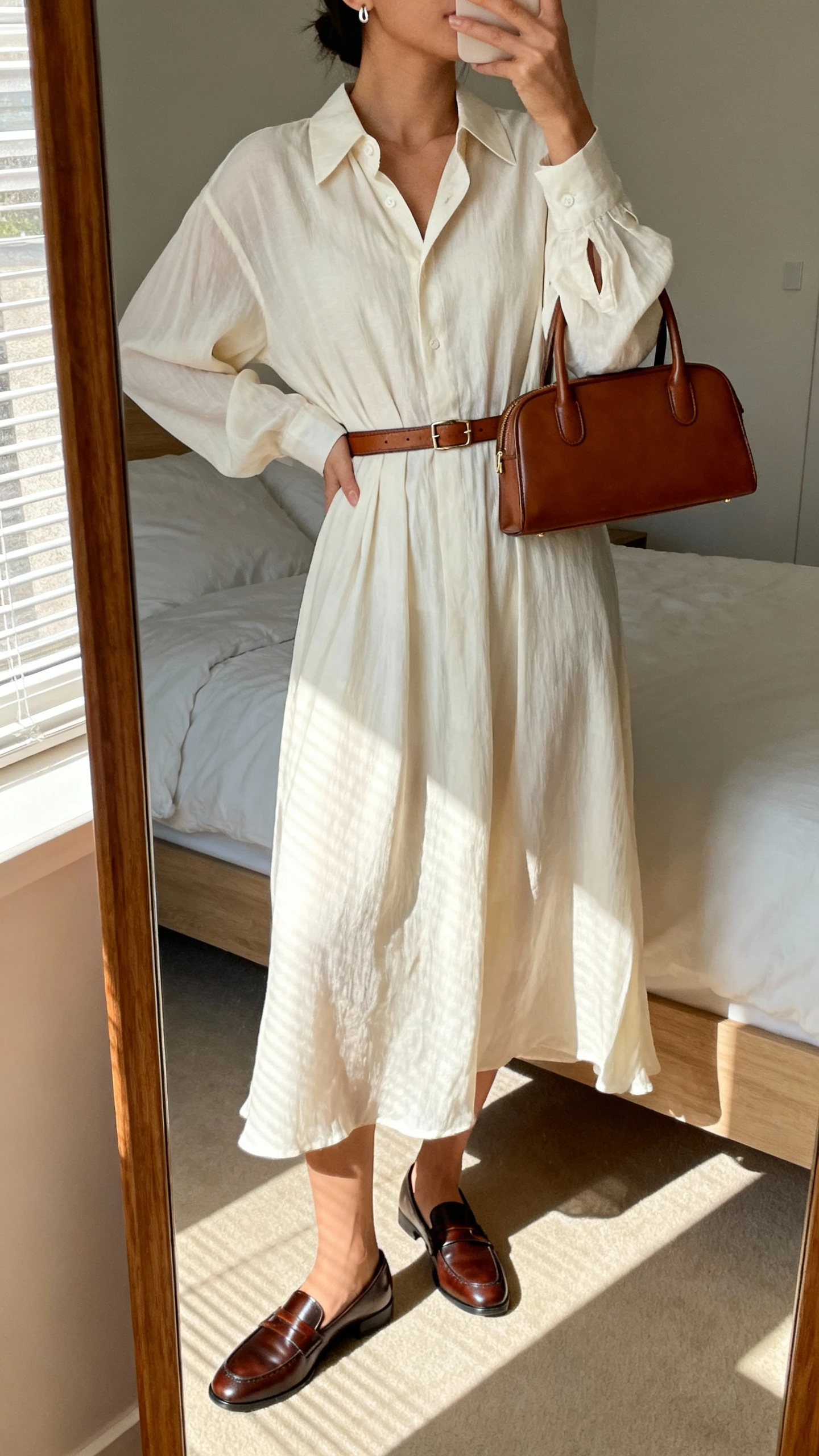 iPhone mirror selfie of a woman wearing a flowing midi shirt dress with a slim leather belt, polished loafers, structured top-handle bag, and minimal stud earrings, face not visible, cropped at neck, bedroom mirror, morning window light, iPhone photo quality.