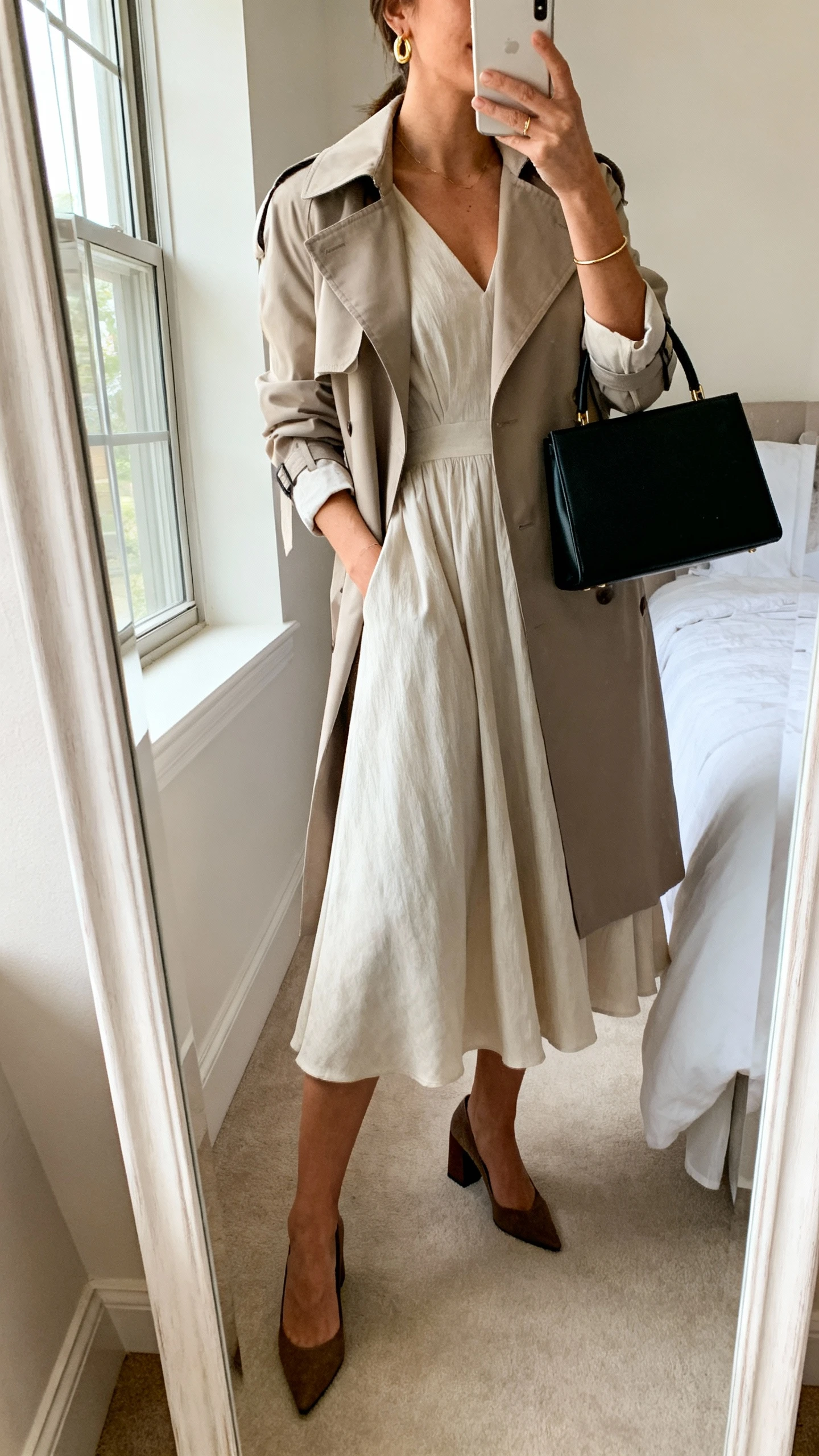 iPhone mirror selfie of a woman wearing a flowy A-line midi dress with elbow-length sleeves, a lightweight trench, block-heel pumps, a structured top-handle bag in black, delicate gold earrings, and a slim bracelet; face not visible, cropped at neck, bedroom full-length mirror, natural morning window light, iPhone photo quality.
