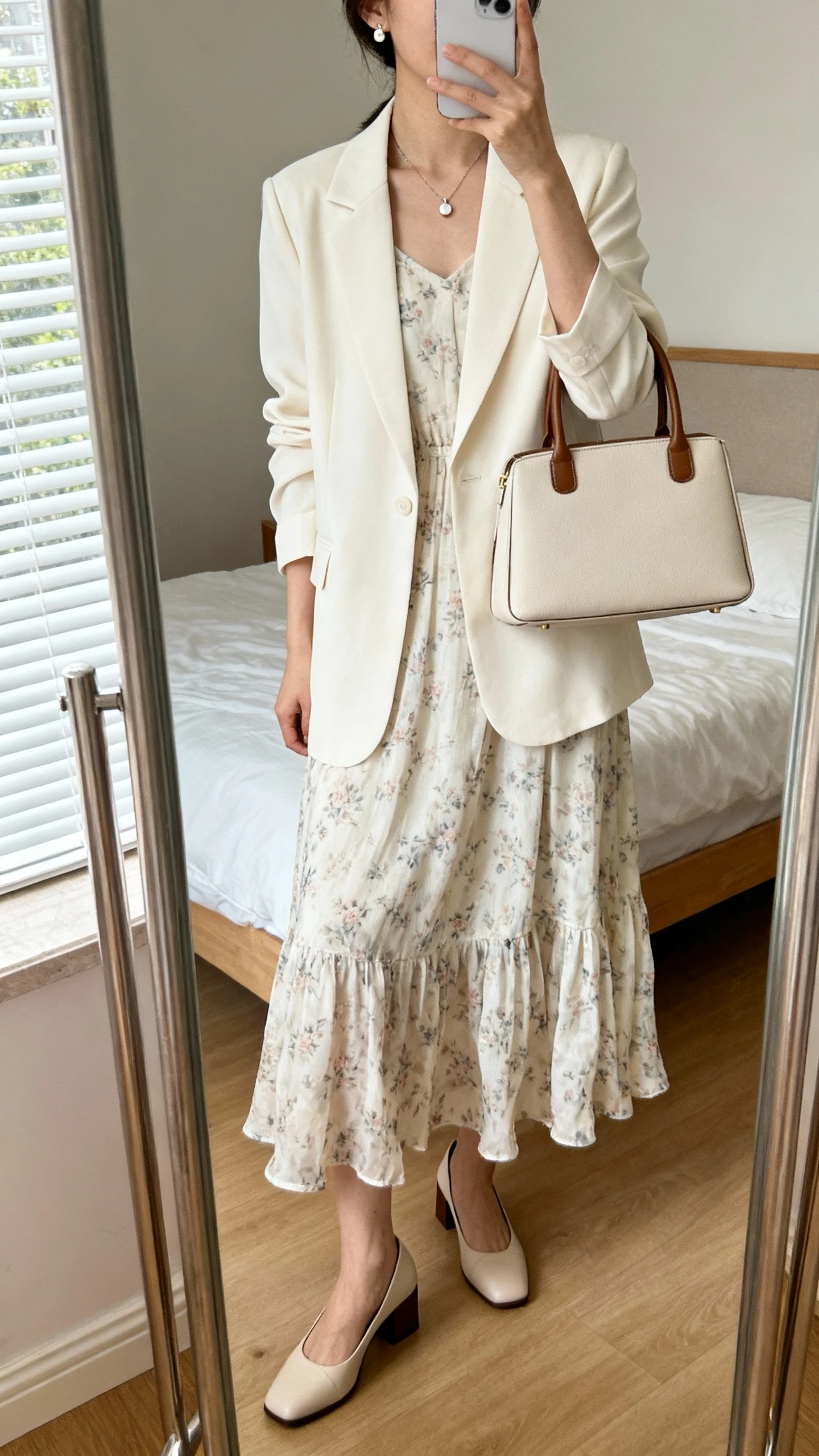 iPhone mirror selfie of a woman wearing a flowy midi dress in a subtle floral print with a cream tailored blazer, closed-toe block heels, a delicate pendant necklace, small stud earrings, and a structured top-handle bag, face not visible, cropped at neck, bedroom mirror with natural window light, iPhone photo quality.