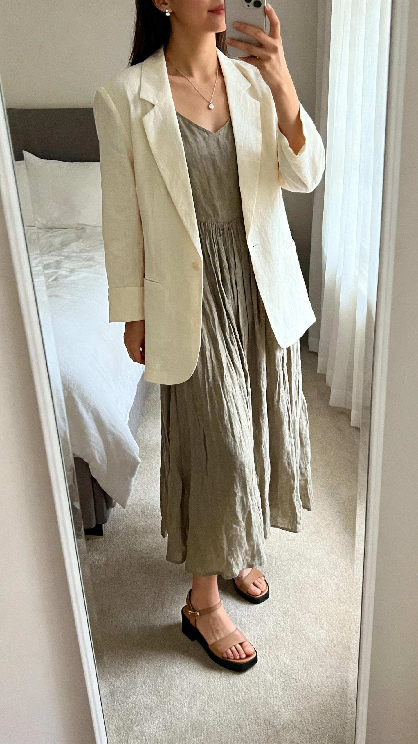 iPhone mirror selfie of a woman wearing a flowy midi dress in breathable cotton with a modest neckline, a soft unlined blazer in cream, low block-heel sandals, and a delicate pendant necklace with small stud earrings, face not visible, cropped at neck, bedroom full-length mirror, morning window light, iPhone photo quality.