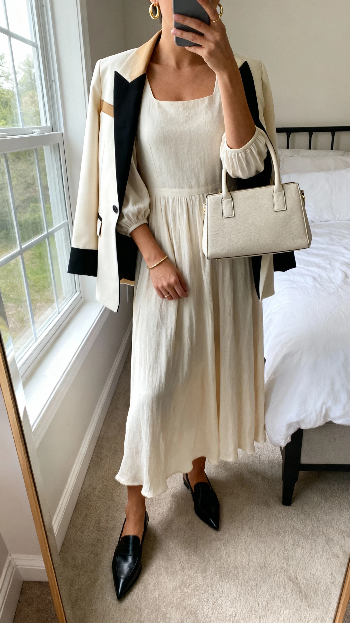 iPhone mirror selfie of a woman wearing a flowy midi dress with a crew or square neckline and elbow-length sleeves, a tailored blazer in cream/tan/black, pointed-toe low heels or sleek loafers, a structured top-handle bag, delicate gold hoop earrings, and a thin bracelet; face not visible, cropped at neck, bedroom mirror, natural window light, iPhone photo quality.