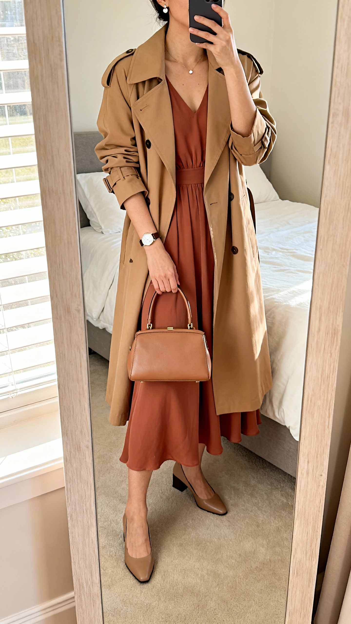 iPhone mirror selfie of a woman wearing a flowy solid-color midi dress with elbow-length sleeves, a lightweight tan trench coat, low block-heel pumps, a structured top-handle bag, delicate stud earrings, and a simple watch; face not visible, cropped at neck, bedroom full-length mirror, soft morning window light, iPhone photo quality.