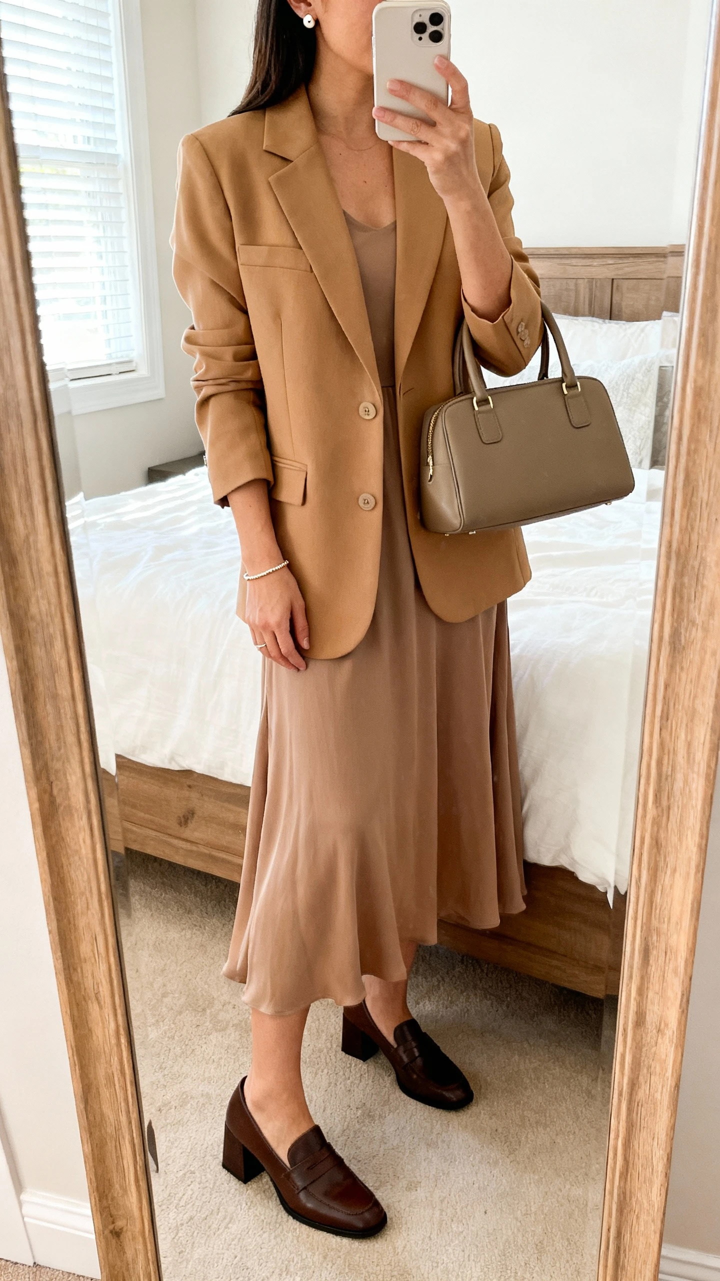 iPhone mirror selfie of a woman wearing a flowy solid-color midi dress, a tailored camel blazer, block-heel loafers, a structured top-handle bag, simple stud earrings, and a delicate bracelet; face not visible, cropped at neck, bedroom mirror with morning window light, iPhone photo quality.