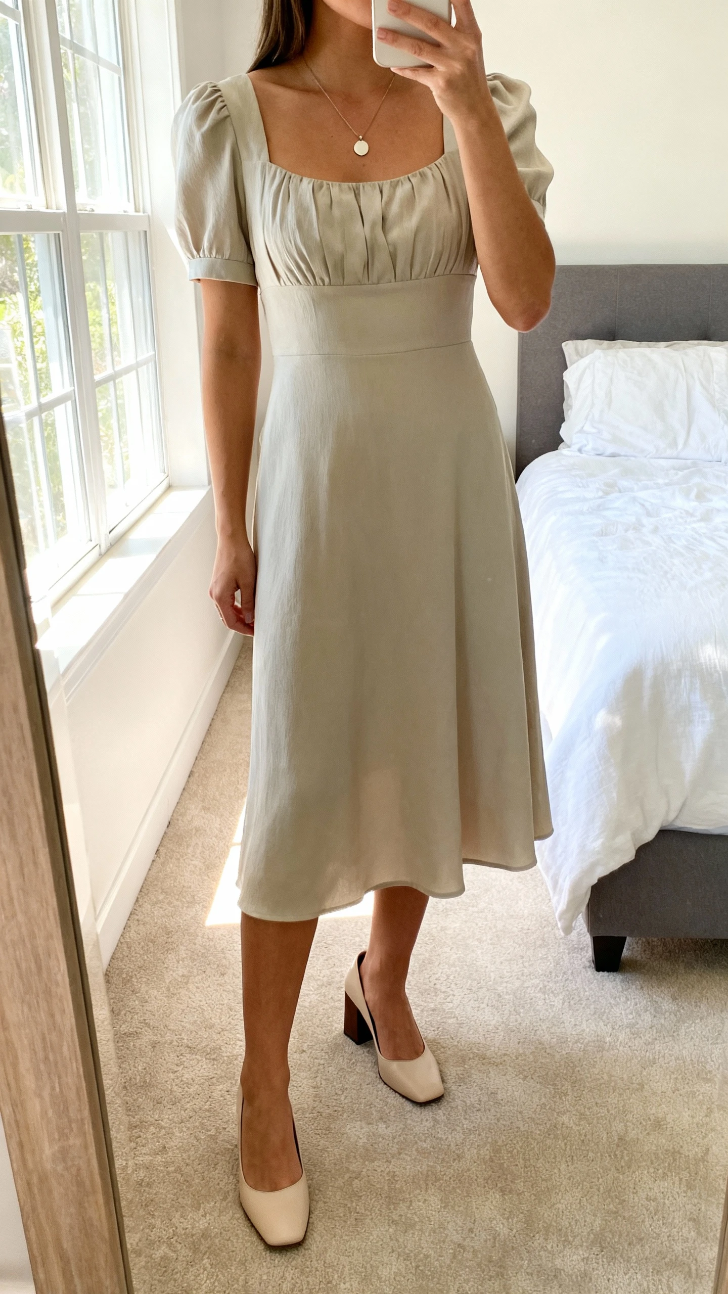 iPhone mirror selfie of a woman wearing a graceful midi dress with a softly structured bodice and modest hem, paired with closed-toe block heels and a simple pendant necklace, face not visible, cropped at neck, bedroom mirror, natural window light, iPhone photo quality.