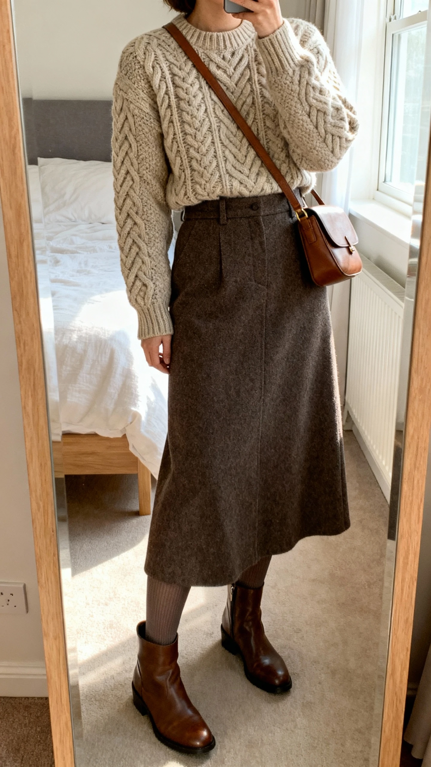 iPhone mirror selfie of a woman wearing a heritage cable-knit sweater, high-waisted wool maxi skirt, opaque tights, leather ankle boots, and a simple leather crossbody, face not visible, cropped at neck, bedroom mirror with natural morning window light, iPhone photo quality