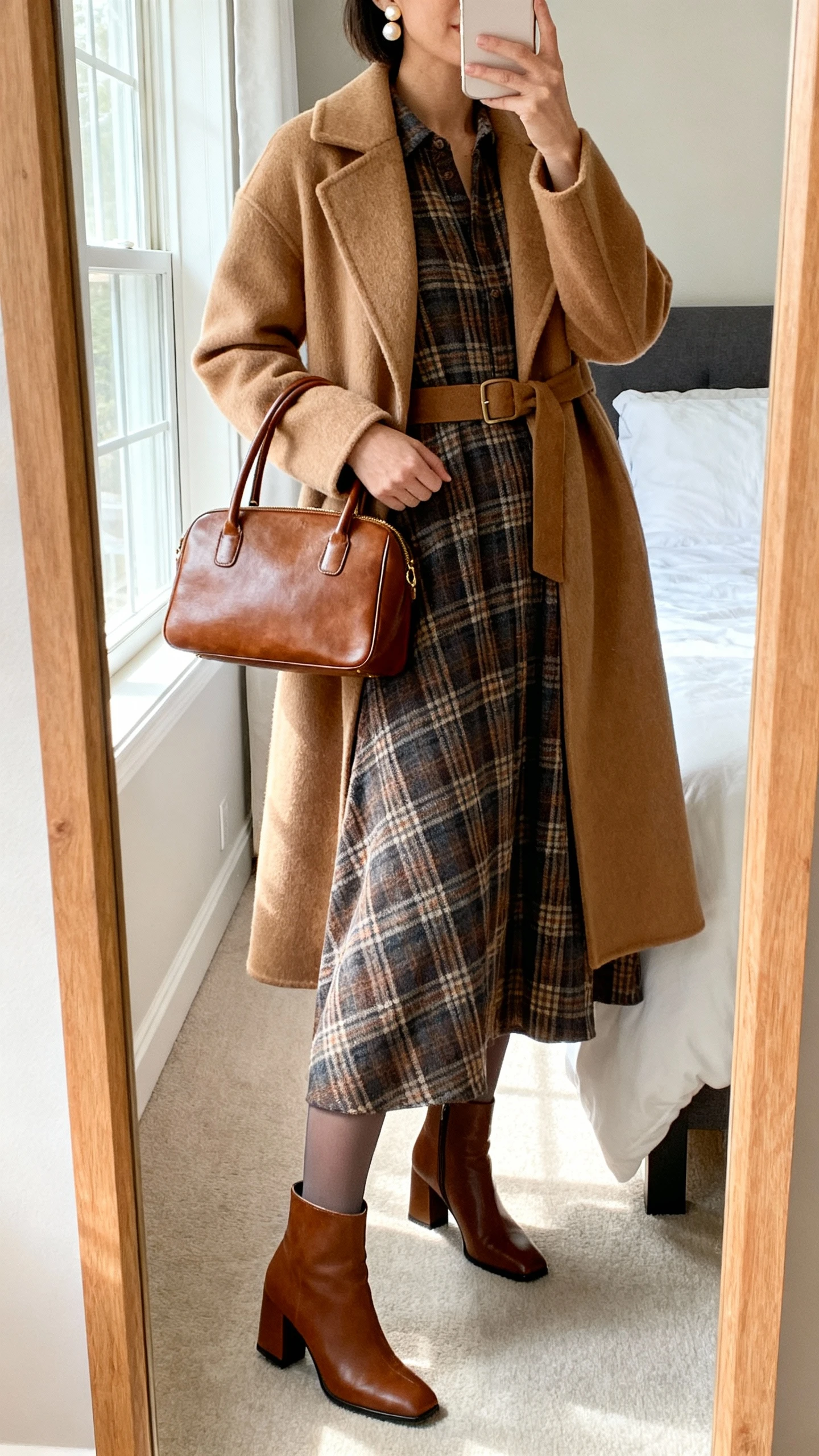 iPhone mirror selfie of a woman wearing a heritage plaid maxi dress styled with a belted camel wool coat, opaque tights, block-heel ankle boots, structured leather top-handle bag, and pearl stud earrings, face not visible, cropped at neck, bedroom mirror, morning window light, iPhone photo quality.