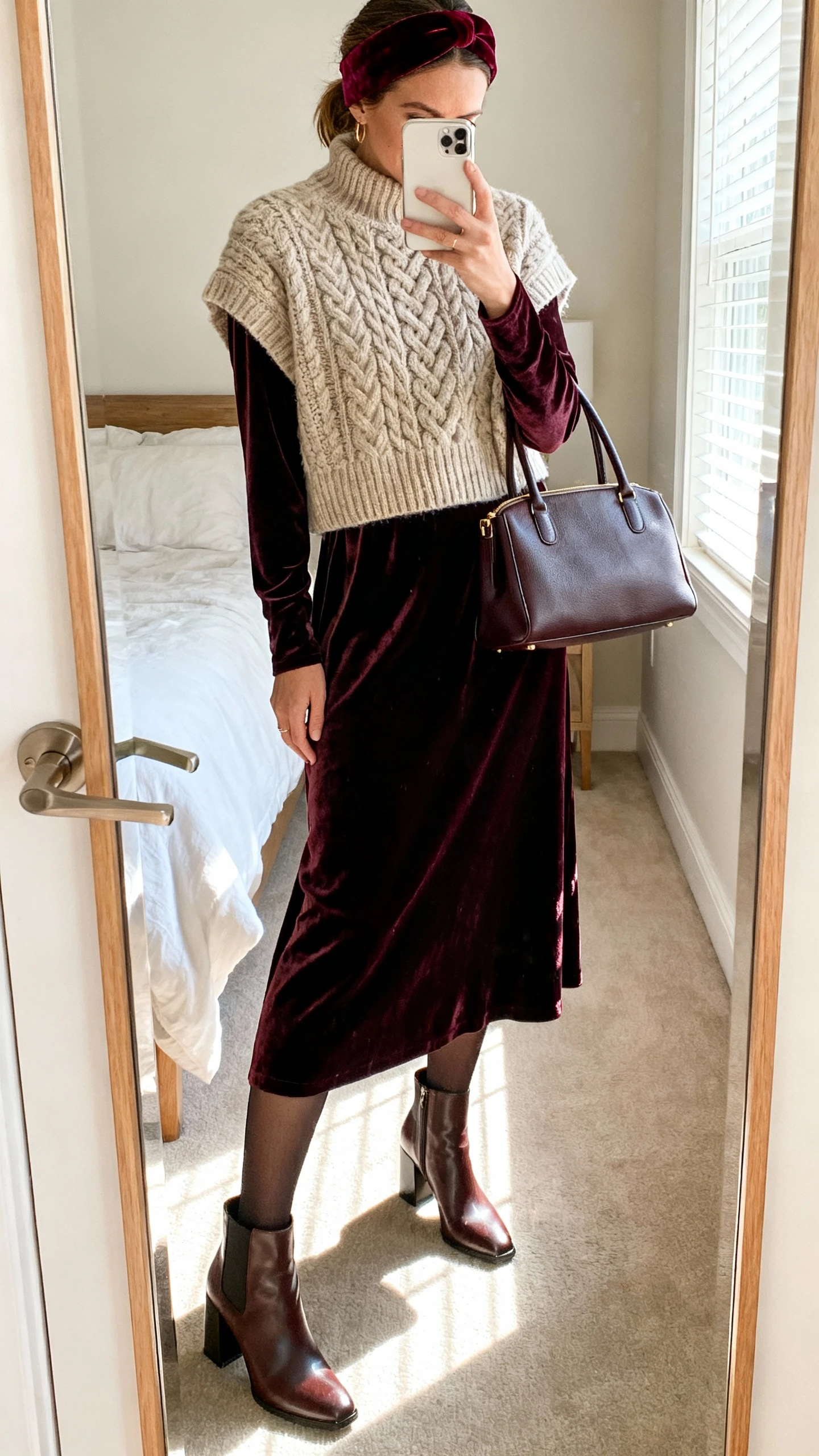 iPhone mirror selfie of a woman wearing a heritage cable-knit sweater layered over a long-sleeve velvet maxi dress, opaque tights, block-heel ankle boots, a velvet headband, and a structured top-handle bag, face not visible, cropped at neck, bedroom mirror with morning window light, iPhone photo quality.
