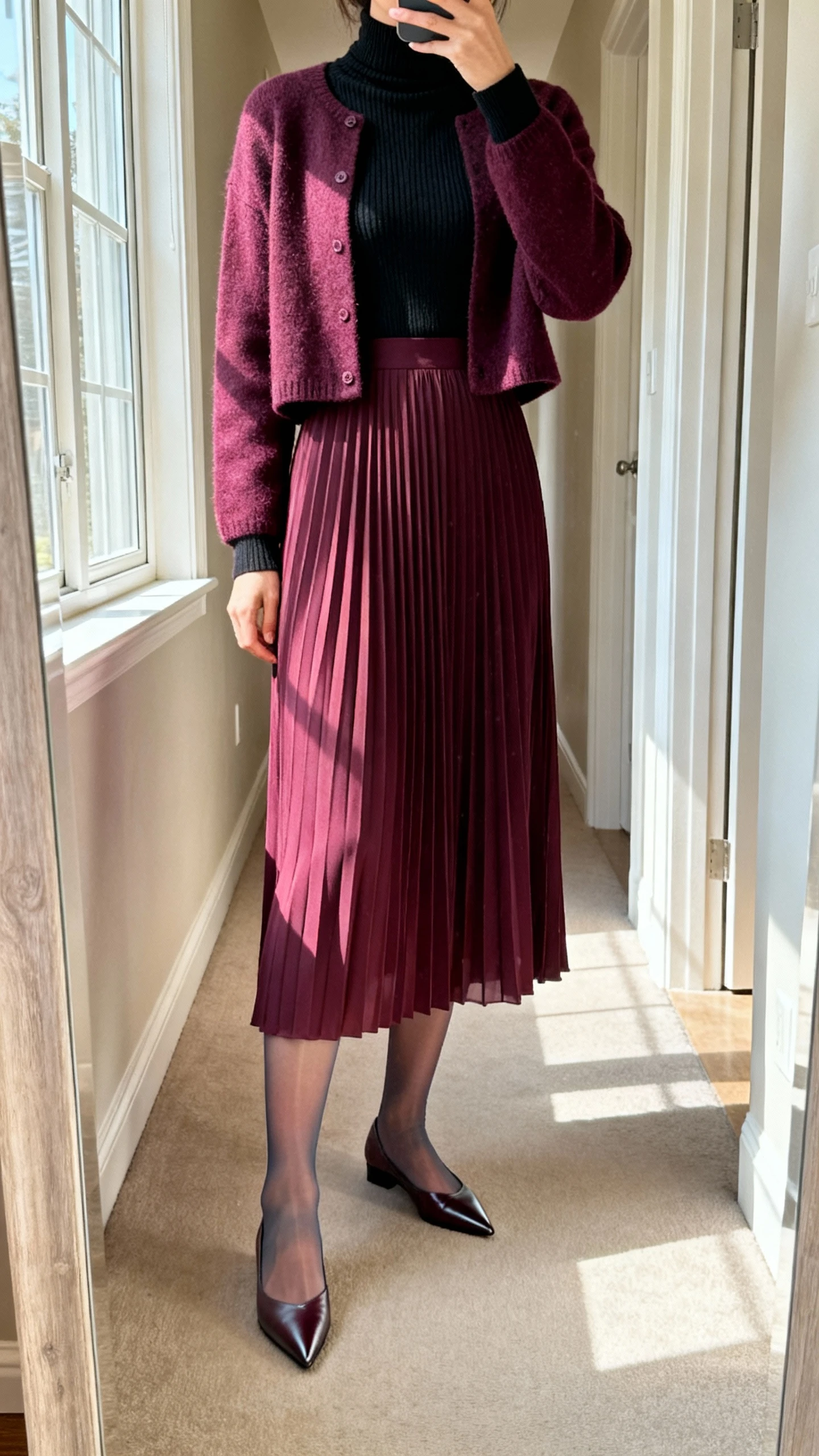 iPhone mirror selfie of a woman wearing a jewel-tone long-sleeve pleated midi dress over a fitted black merino turtleneck, topped with a cropped cardigan, with pointed-toe flats and sheer-to-opaque tights; face not visible, cropped at shoulders, full-length mirror in hallway, morning window light, iPhone photo quality.