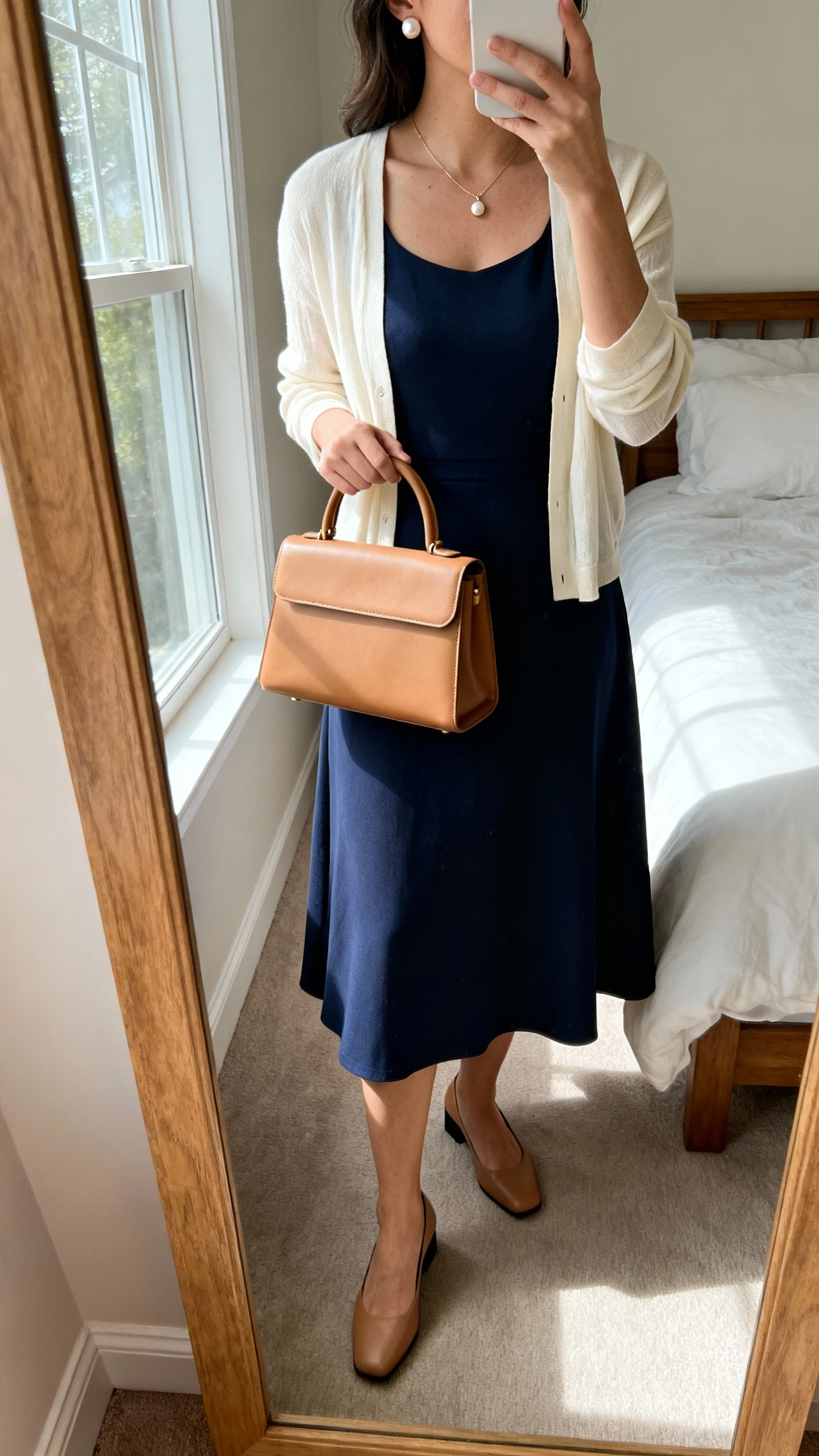 iPhone mirror selfie of a woman wearing a knee-length fit-and-flare dress in navy with a lightweight cream cardigan, low block heels, a structured top-handle bag in tan, pearl stud earrings, and a delicate pendant necklace; face not visible, cropped at neck, bedroom mirror, natural window light, iPhone photo quality.
