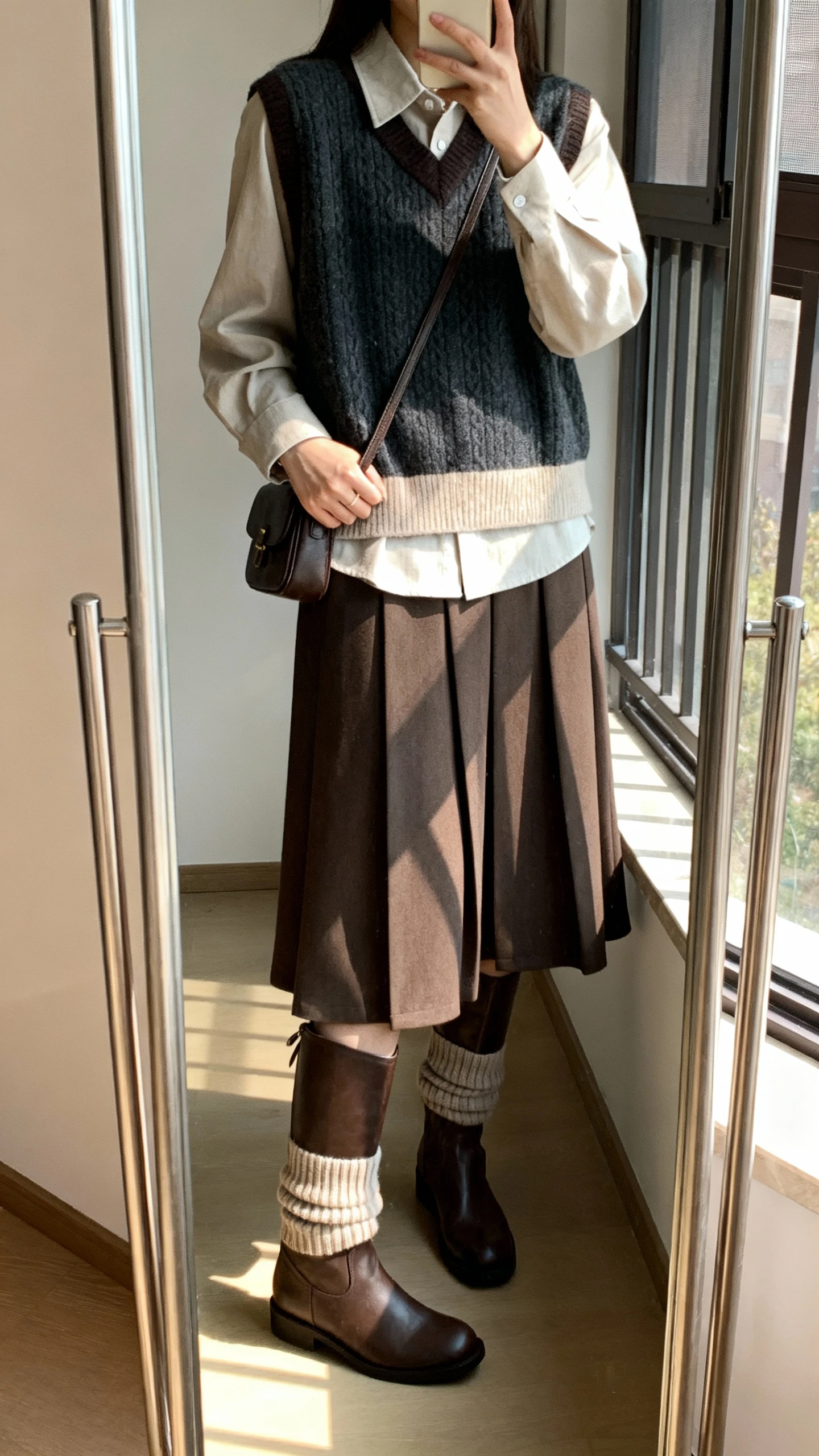 iPhone mirror selfie of a woman wearing a layered knit vest over a collared midi dress with knee-high boots, ribbed socks peeking, and a small satchel, face not visible, cropped at shoulders, full-length mirror by window, gentle morning light, iPhone photo quality.