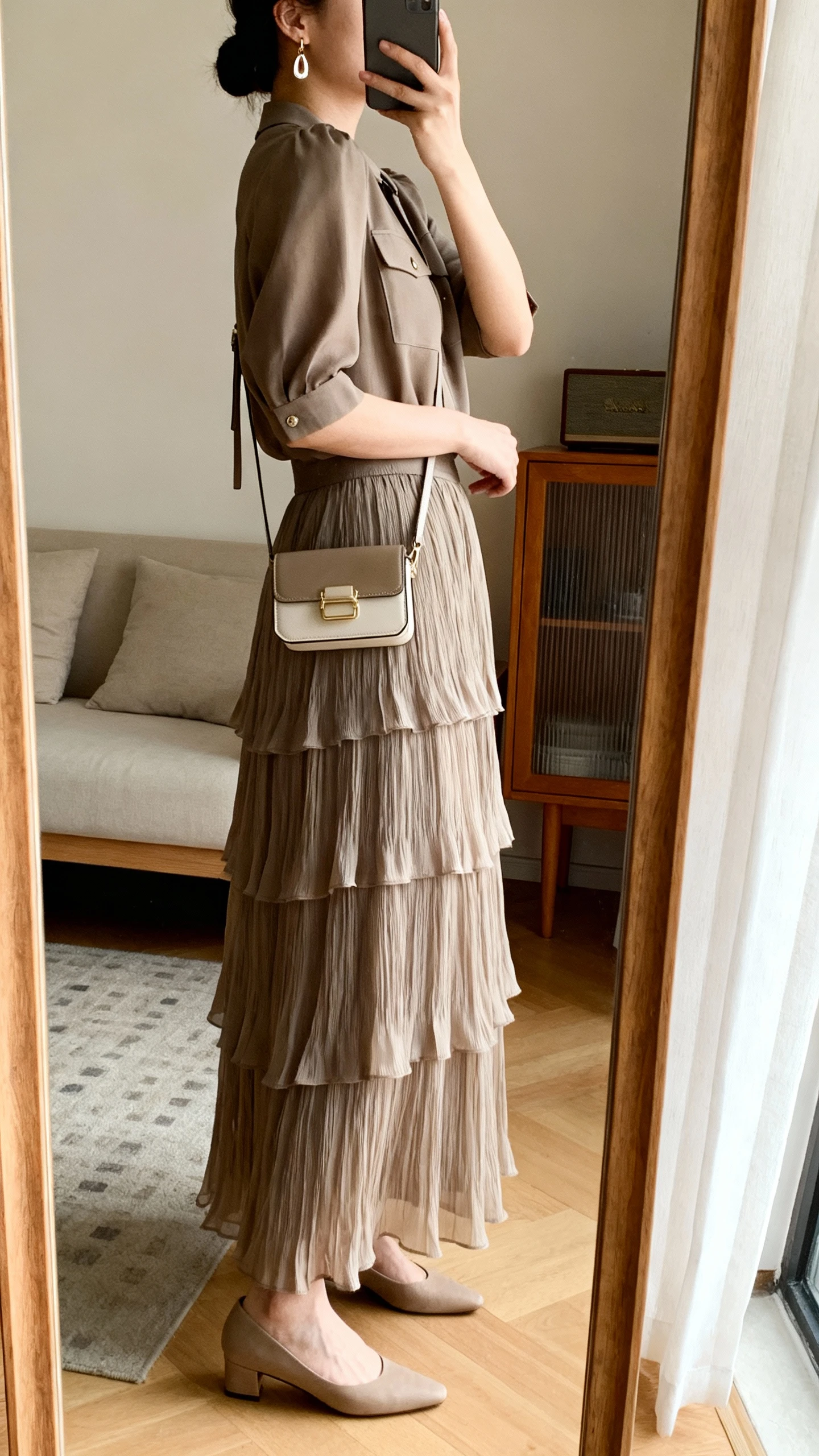 iPhone mirror selfie of a woman wearing a layered maxi skirt with a structured top, low heels, subtle drop earrings, and a compact crossbody bag, face not visible, side angle, living room full-length mirror, warm indoor lighting, iPhone photo quality.