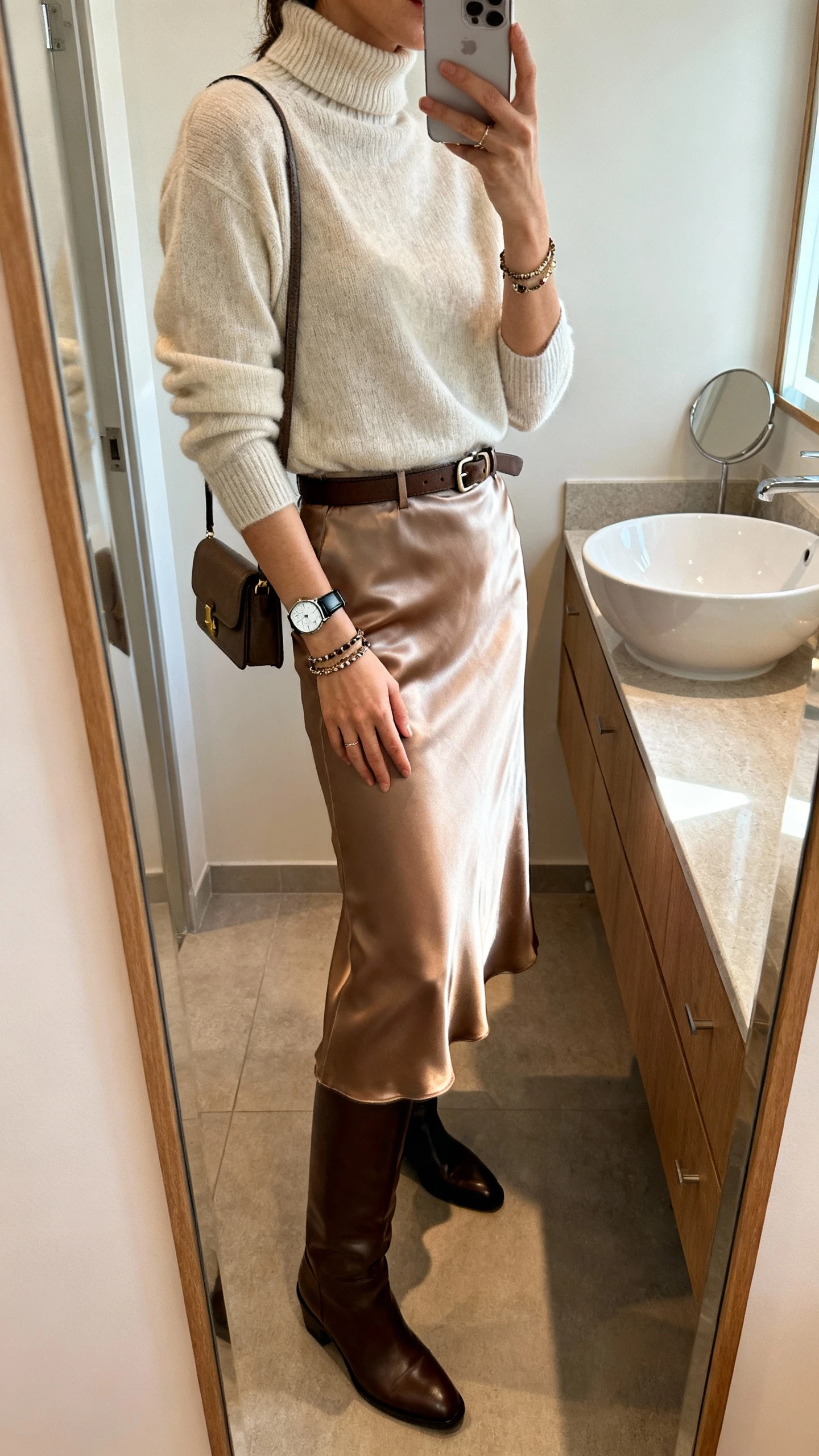 iPhone mirror selfie of a woman wearing a lightweight mock-neck sweater tucked into a satin midi skirt with knee-high boots, layered bracelets and a sleek watch, and a small shoulder bag (optional waist belt), face not visible, side angle, bathroom mirror, soft indoor morning light, iPhone photo quality.