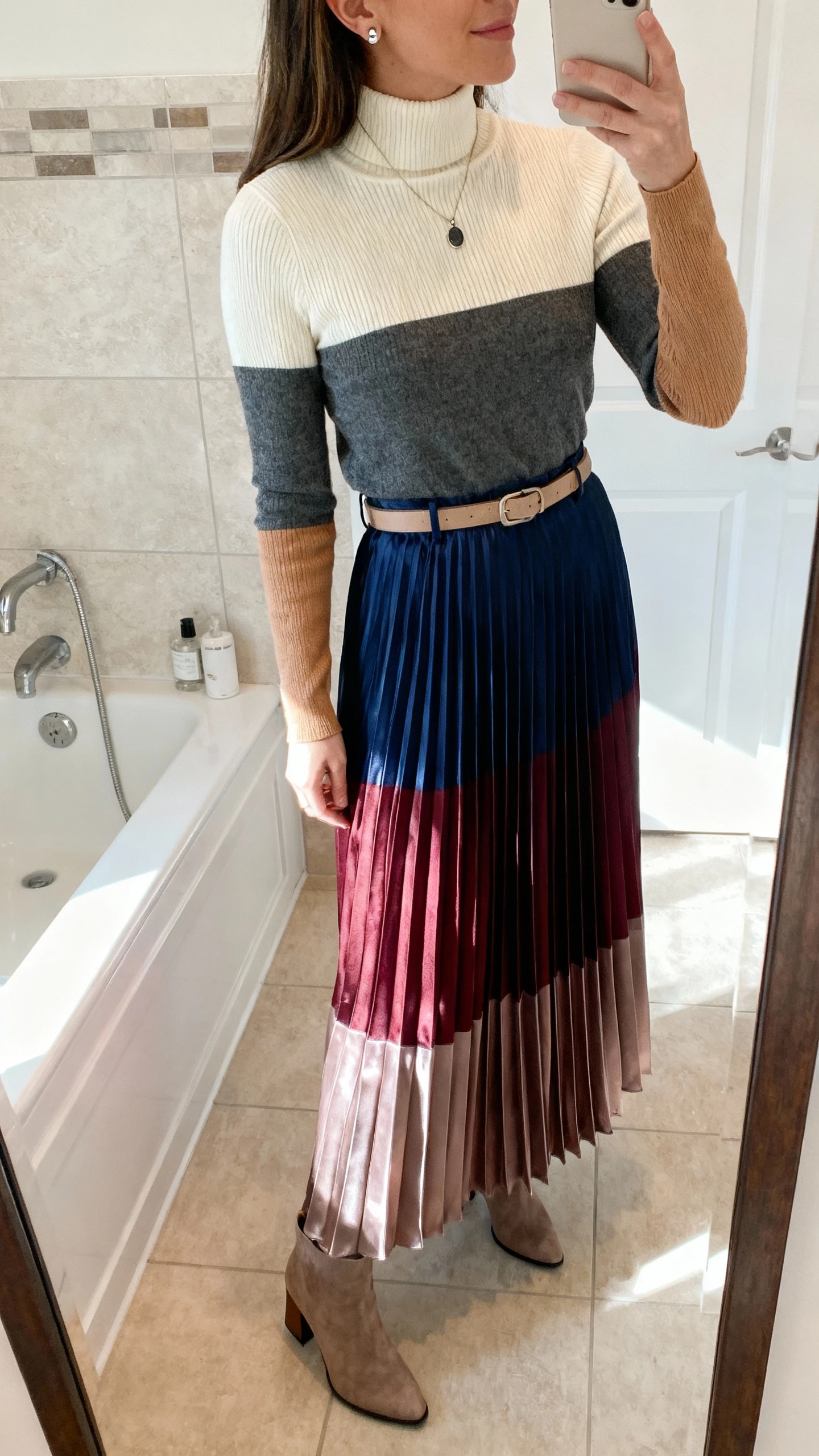 iPhone mirror selfie of a woman wearing a lightweight ribbed turtleneck (cream, charcoal, or camel), a pleated maxi skirt in satin or chiffon (navy, burgundy, or soft metallic), classic ankle boots or block-heel pumps, an optional minimalist belt, and simple stud earrings with a pendant necklace; face not visible, side angle, bathroom mirror, morning light, iPhone photo quality.