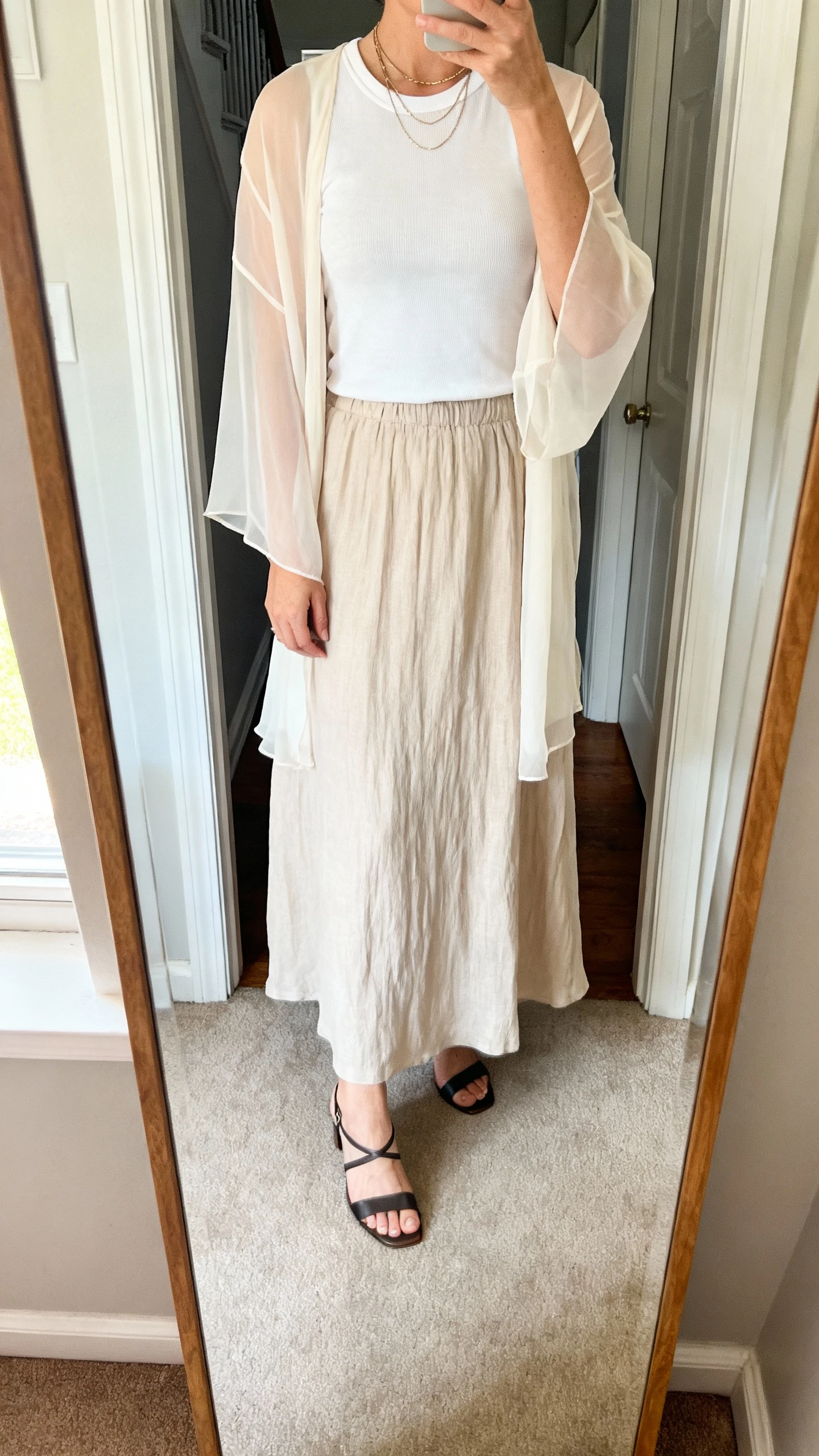 iPhone mirror selfie of a woman wearing a lightweight maxi skirt (cotton voile/crepe/rayon, solid or subtle print) with a fitted tee or knit shell (high crew or boat neckline), a sheer cardigan or chiffon topper covering shoulders and upper arms, strappy low heels or sleek flat sandals, and layered delicate necklaces; face not visible, cropped at neck, entryway full-length mirror, natural morning light, iPhone photo quality.