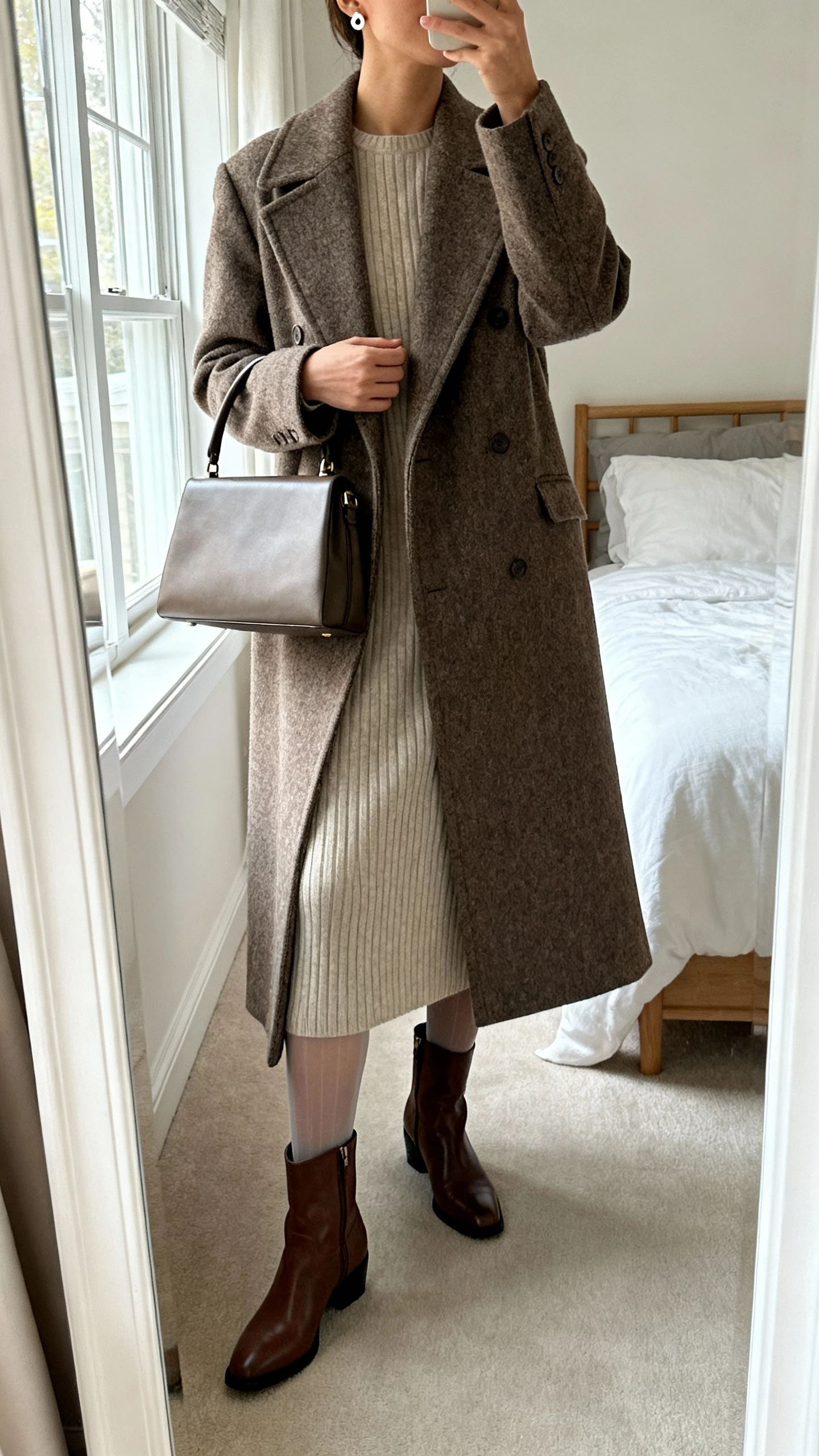 iPhone mirror selfie of a woman wearing a long heritage wool coat over a ribbed knit midi dress, sheer tights, classic leather ankle boots, structured top-handle bag, and delicate stud earrings, face not visible, cropped at neck, bedroom mirror with morning window light, iPhone photo quality.