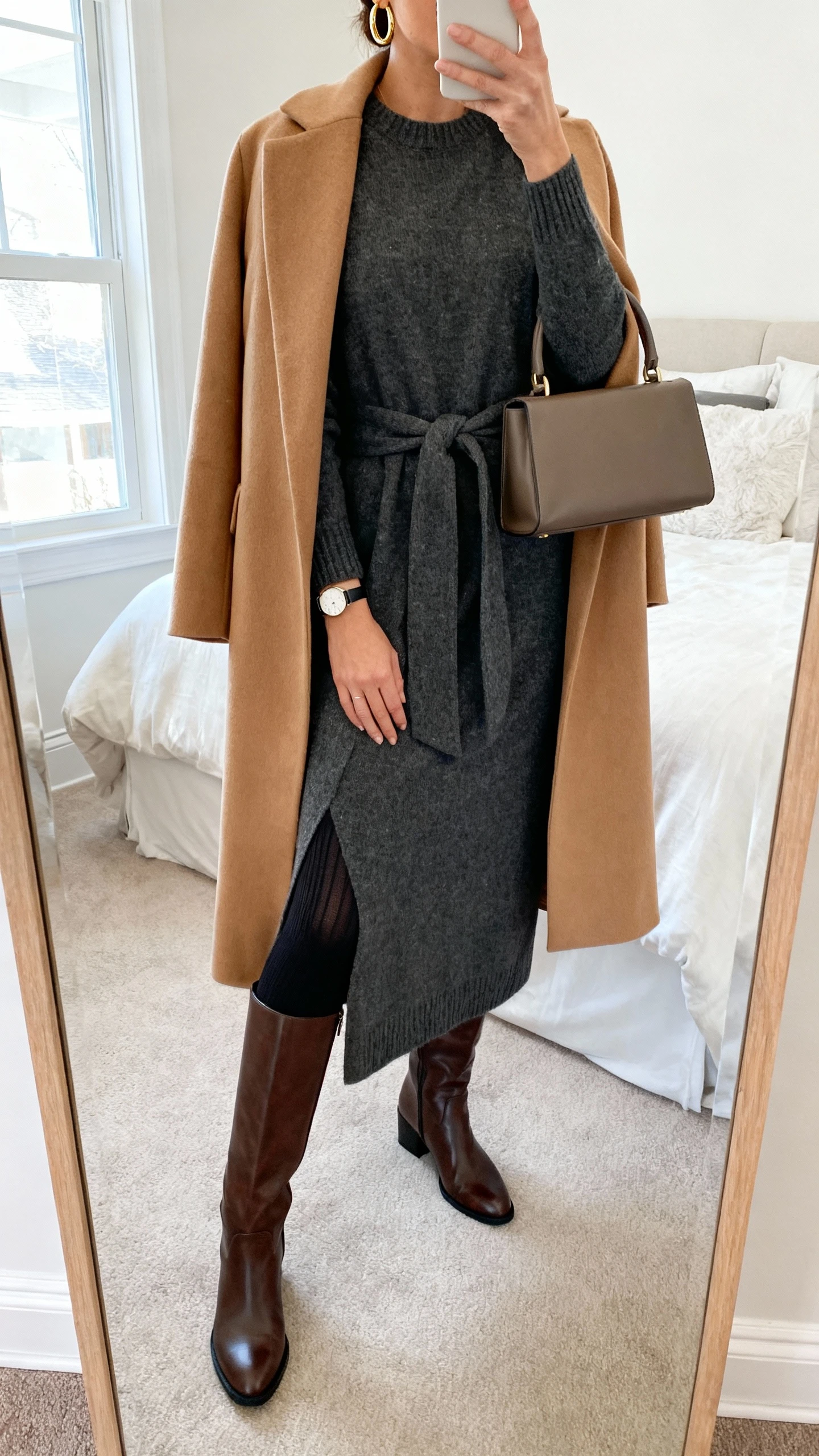 iPhone mirror selfie of a woman wearing a long-sleeve wool-blend knit maxi dress in charcoal with a tailored camel wrap coat, leather knee-high boots with a low block heel, opaque black thermal tights, simple gold hoop earrings, a slim watch, and a structured top-handle bag; face not visible, cropped at neck, bedroom mirror, natural window light, iPhone photo quality.