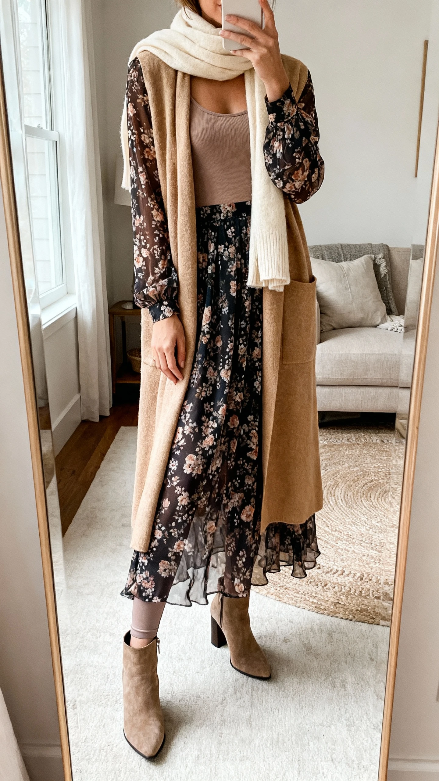 iPhone mirror selfie of a woman wearing a long-sleeve deep floral chiffon maxi dress with a thermal camisole and thin leggings underneath, a sand longline cardigan coat, block-heel boots, and a soft cream scarf; face not visible, cropped at neck, living room mirror, natural daylight, iPhone photo quality.