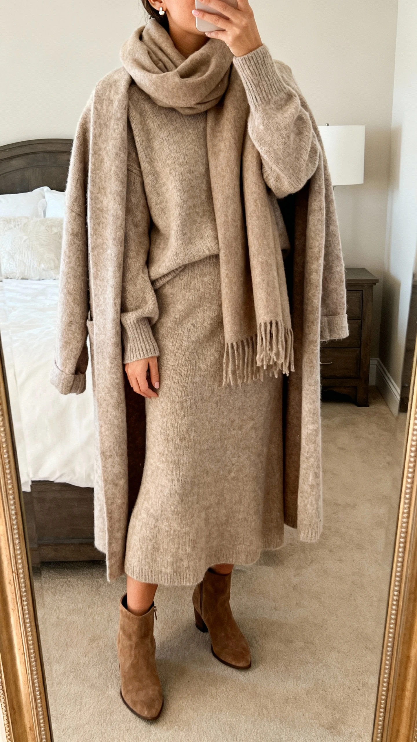 iPhone mirror selfie of a woman wearing a luxe matching knit set (relaxed sweater and midi skirt) with an oversized scarf, cozy longline cardigan draped over shoulders, suede ankle boots, and delicate stud earrings, face not visible, cropped at shoulders, bedroom mirror, warm indoor lighting, iPhone photo quality.