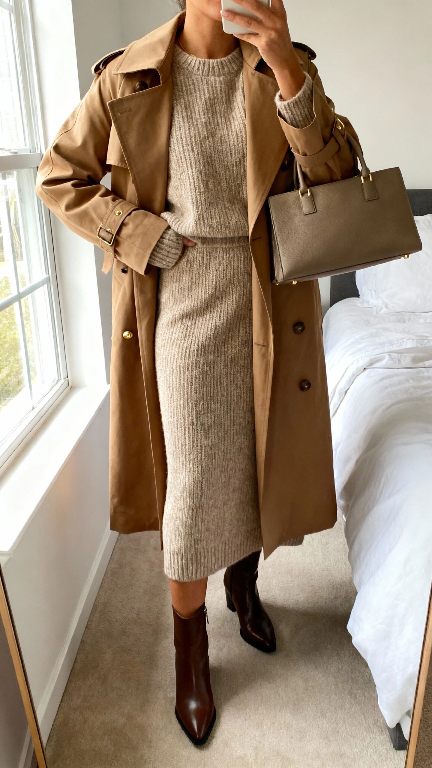 iPhone mirror selfie of a woman wearing a matching knit sweater and knit midi skirt set with a classic camel trench coat, sleek leather ankle boots, minimal gold studs, and a structured tote, face not visible, cropped at neck, bedroom mirror, soft morning window light, iPhone photo quality.