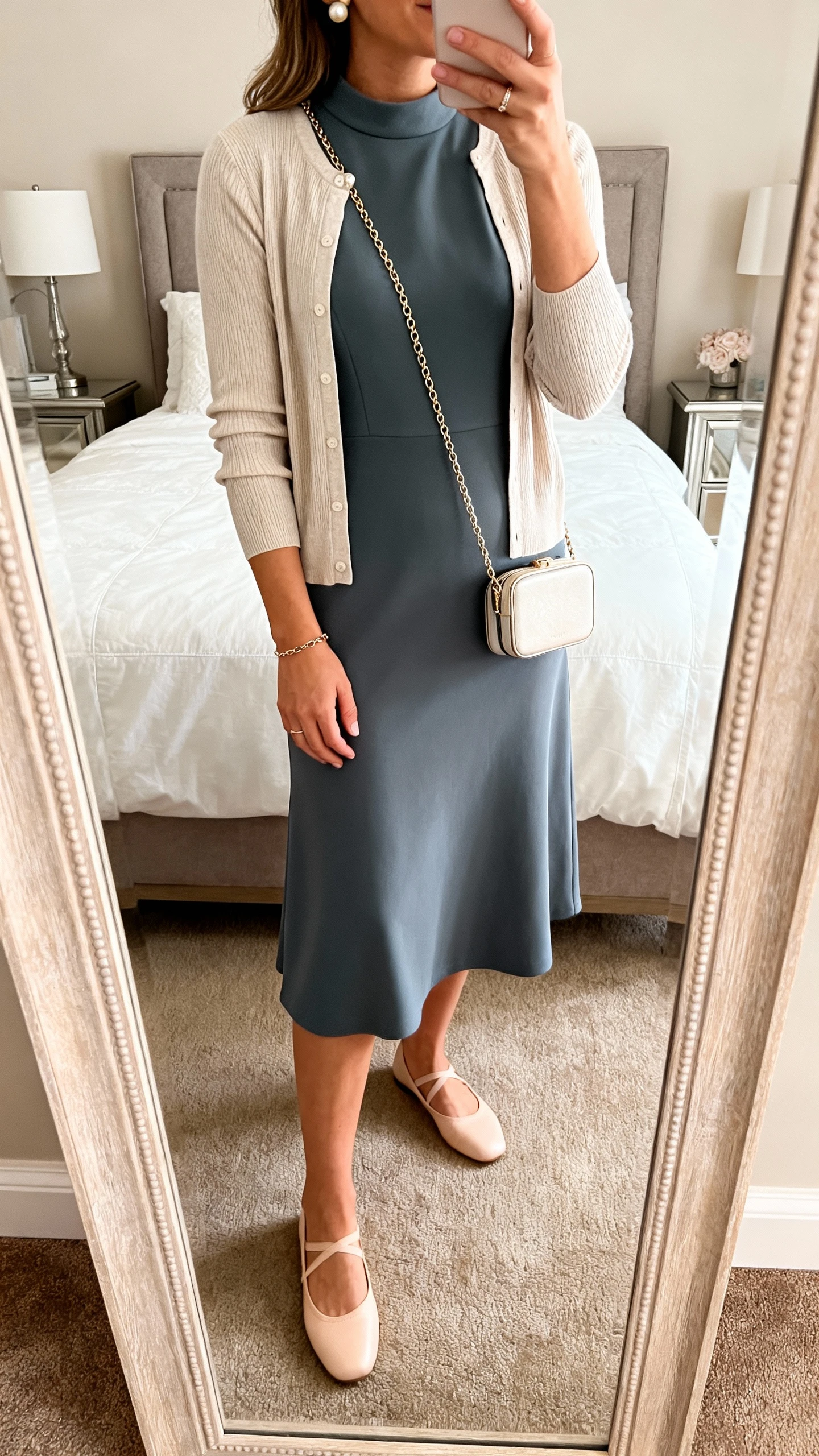 iPhone mirror selfie of a woman wearing a midi A-line dress in ponte with a higher neckline, a fine-gauge cardigan, ballet flats, pearl studs with a dainty chain bracelet, and a compact crossbody, face not visible, cropped at shoulders, bedroom mirror, warm indoor lighting, iPhone photo quality.