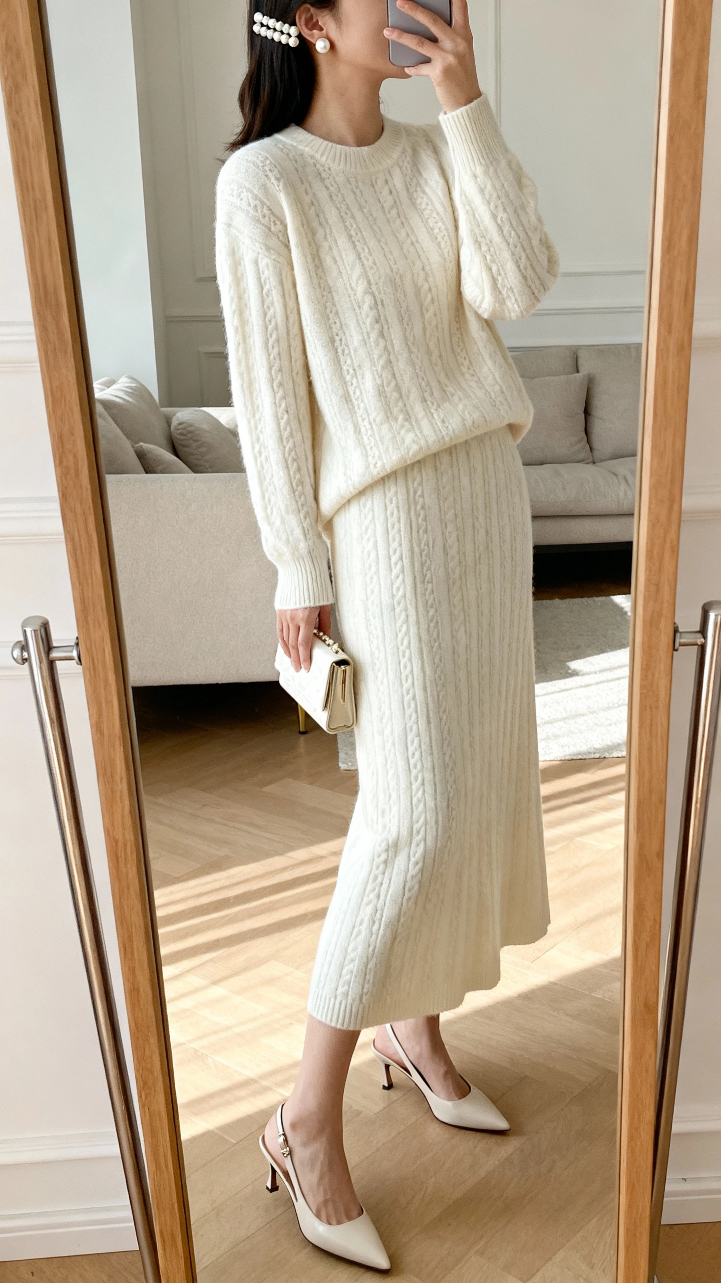 iPhone mirror selfie of a woman wearing a monochrome knit set (matching sweater and midi skirt) with pearl stud earrings, a pearl-accent hair clip, sleek slingback heels, and a small clutch, face not visible, side angle, living room full-length mirror, gentle indoor lighting, iPhone photo quality.
