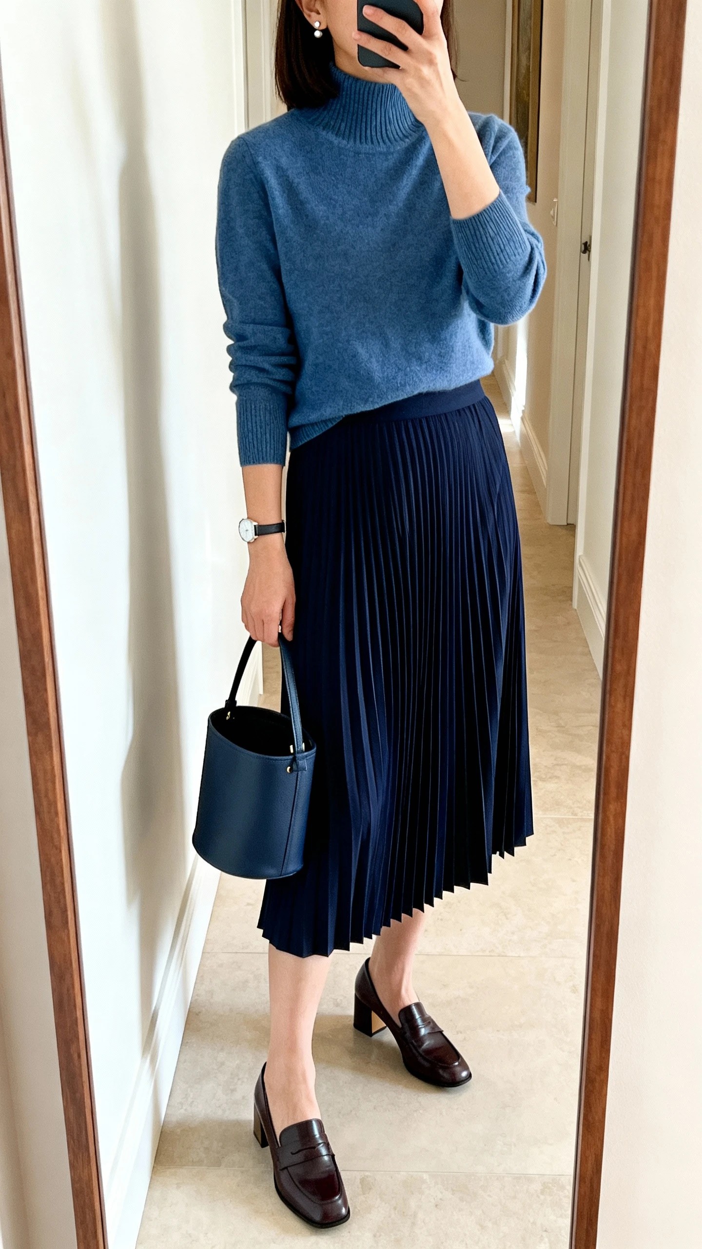 iPhone mirror selfie of a woman wearing a navy midi pleated skirt with a fine-gauge mock-neck knit top tucked in, block-heel loafers, minimalist stud earrings, a thin watch, and a structured bucket bag, face not visible, cropped at shoulders, full-length mirror in hallway, indoor daylight, iPhone photo quality.