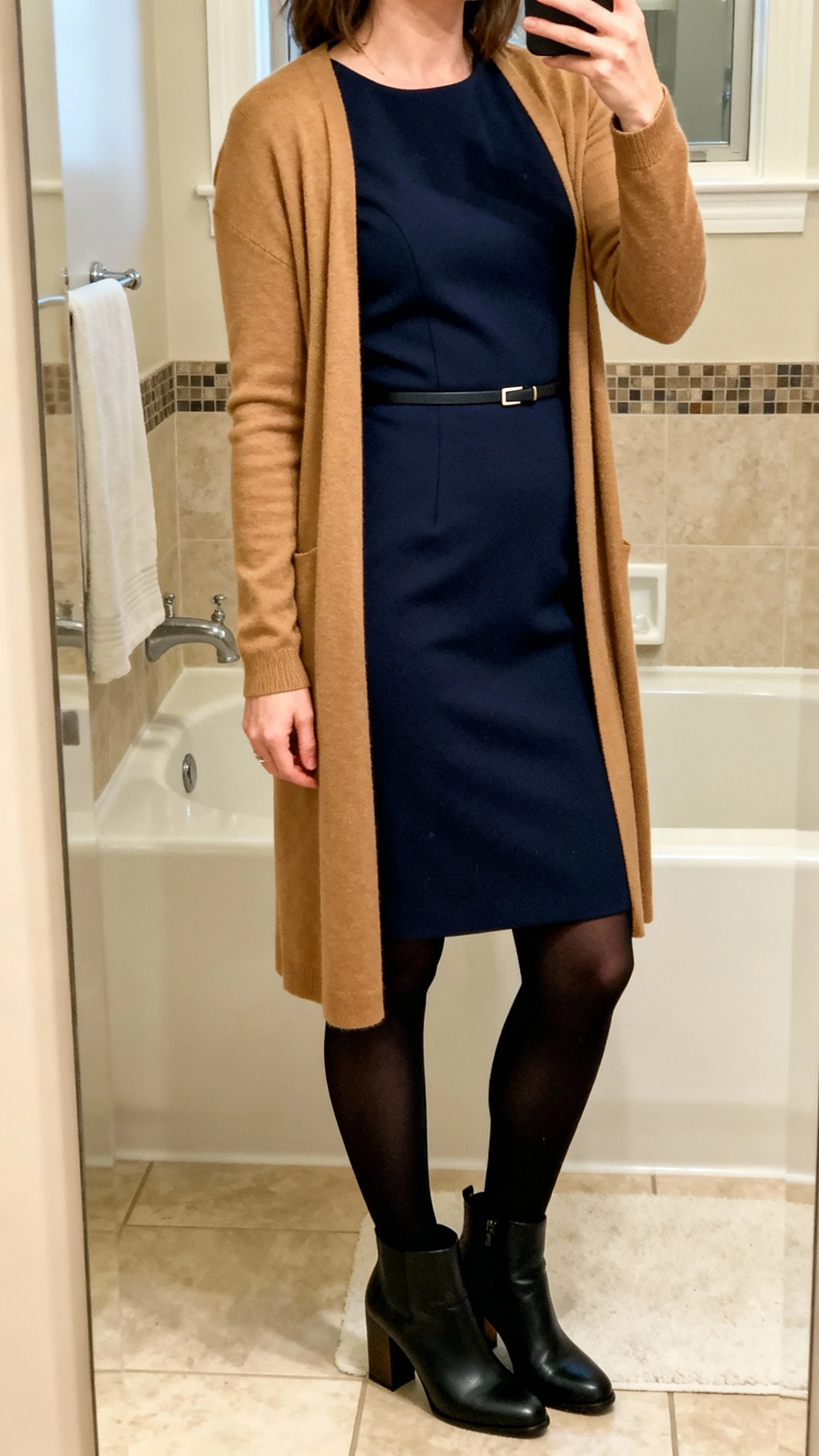 iPhone mirror selfie of a woman wearing a navy midi sheath dress layered with a tan longline cardigan, black opaque tights, block-heel ankle boots, and a minimal waist belt, face not visible, cropped at neck, bathroom mirror with warm indoor lighting, iPhone photo quality.