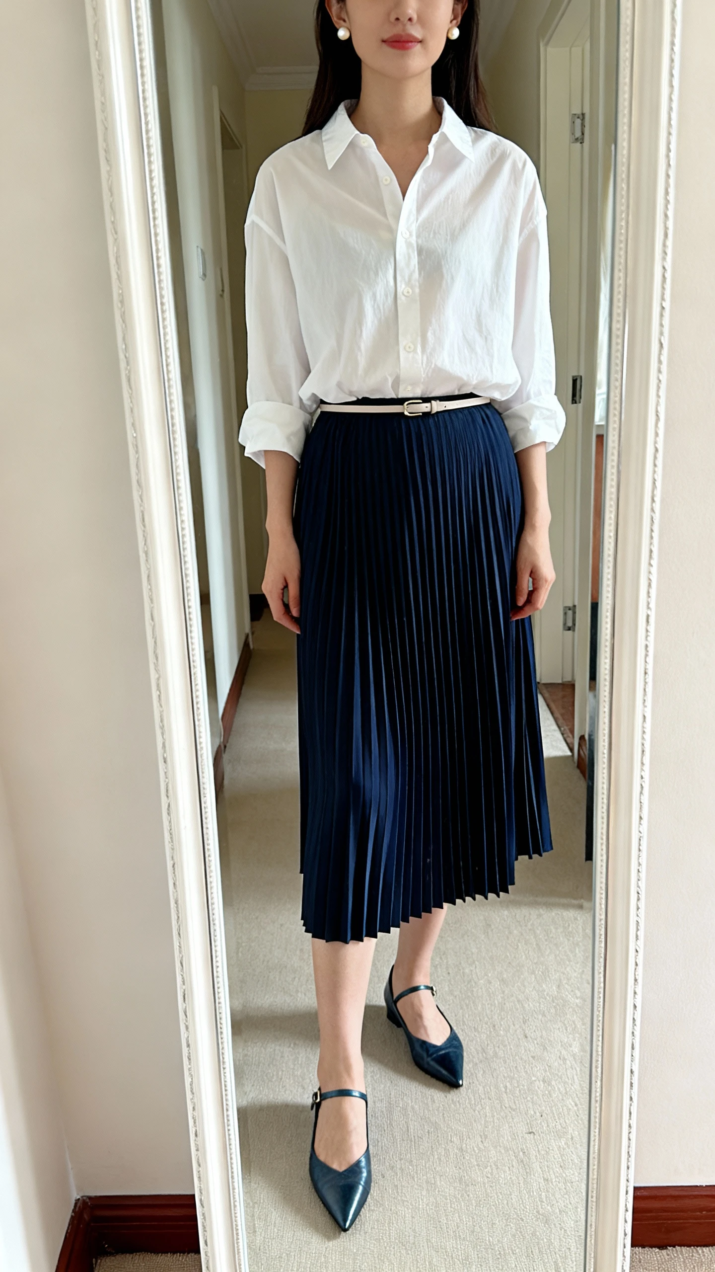iPhone mirror selfie of a woman wearing a navy pleated midi skirt, relaxed white button-up blouse tucked in, pointed-toe flats, a thin belt at the waist, and pearl stud earrings; face not visible, cropped at shoulders, full-length hallway mirror with natural daylight, iPhone photo quality.