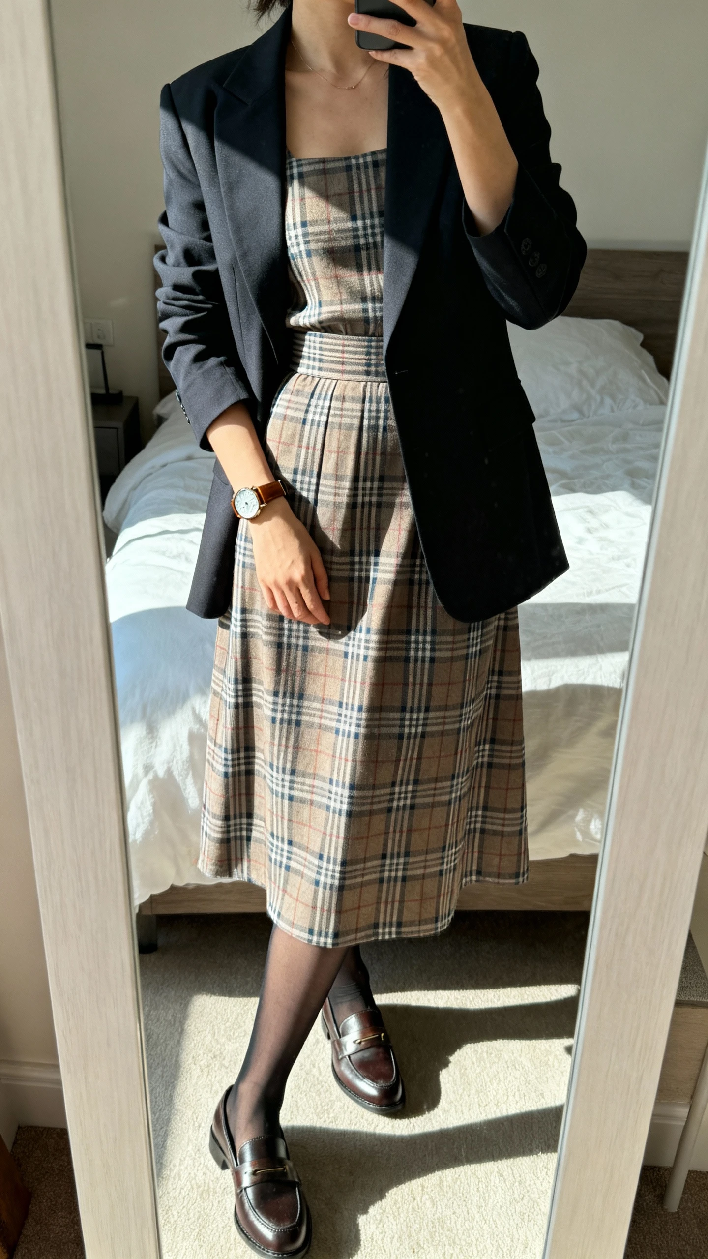 iPhone mirror selfie of a woman wearing a plaid midi dress with a fitted waist, polished blazer, classic loafers, sheer black tights, and a leather watch, face not visible, cropped at shoulders, bedroom mirror, natural daylight, iPhone photo quality.