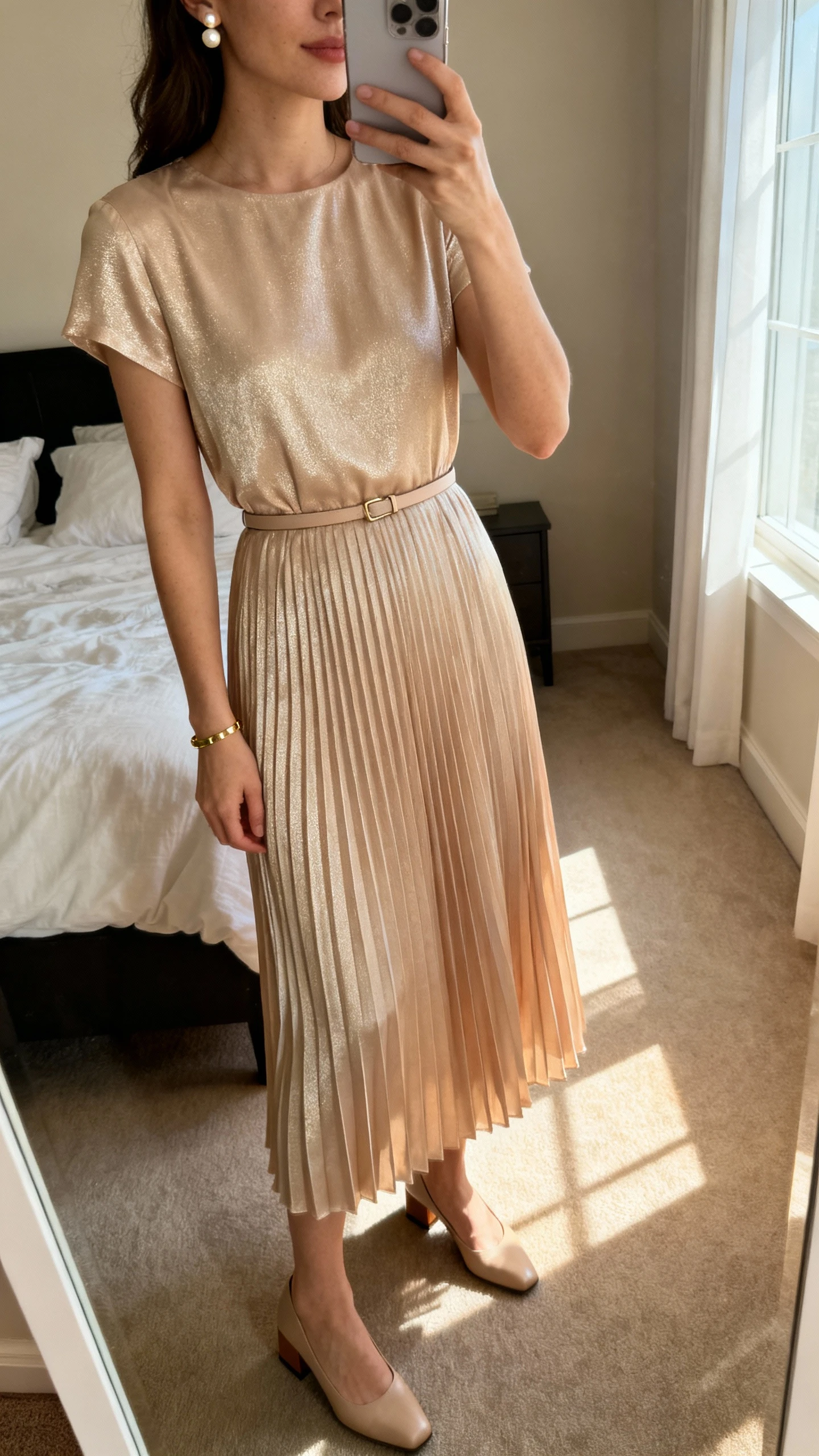 iPhone mirror selfie of a woman wearing a pleated midi dress in soft champagne with a subtle sheen, slim belt at the waist, delicate pearl stud earrings, minimalist gold bracelet, and low block-heel pumps, face not visible, cropped at neck, bedroom mirror with morning window light, iPhone photo quality.