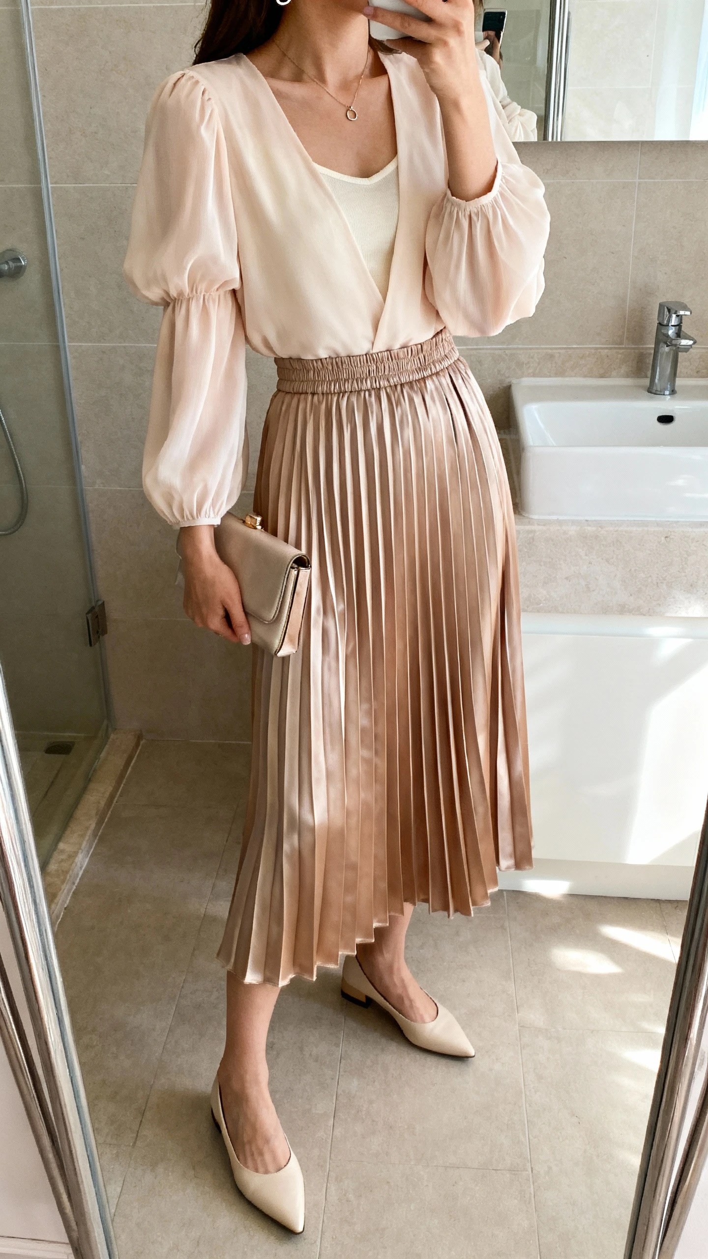 iPhone mirror selfie of a woman wearing a pleated midi skirt in chiffon or satin (elastic or hidden waistband) with a soft blouse with sleeves (puff-sleeve, flutter-sleeve, or long-sleeve chiffon with camisole underneath), pointed-toe flats or low slingback heels, a minimal pendant necklace and small hoops, and a compact clutch; face not visible, side angle, bathroom mirror, morning light, iPhone photo quality.