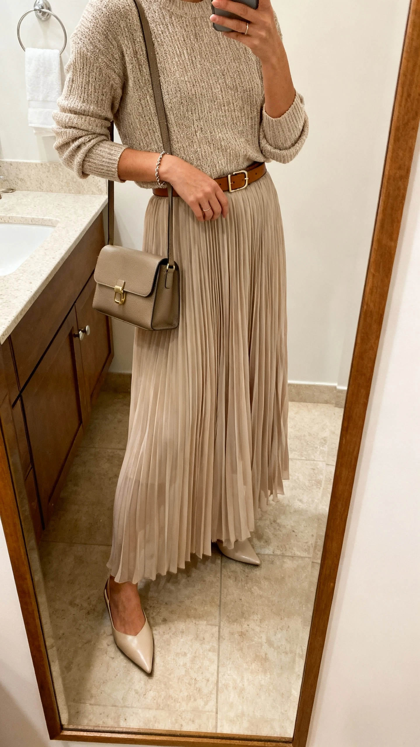 iPhone mirror selfie of a woman wearing a pleated chiffon maxi skirt, a knit top with elbow-length sleeves, pointed-toe flats, a slim leather belt at the waist, and a simple bracelet with a structured small satchel, face not visible, side angle in bathroom mirror, soft indoor lighting, iPhone photo quality.