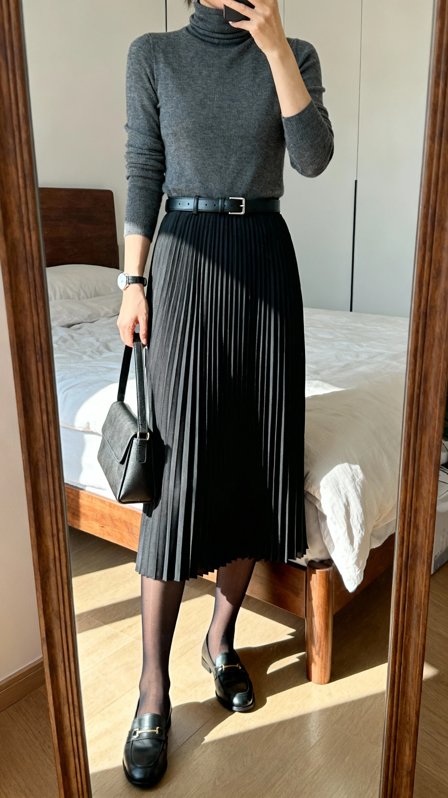 iPhone mirror selfie of a woman wearing a pleated midi skirt with a fitted turtleneck, sheer black tights, loafers, a slim leather belt, and a minimalist watch, holding a structured shoulder bag, face not visible, cropped at shoulders, bedroom mirror, natural daylight, iPhone photo quality.