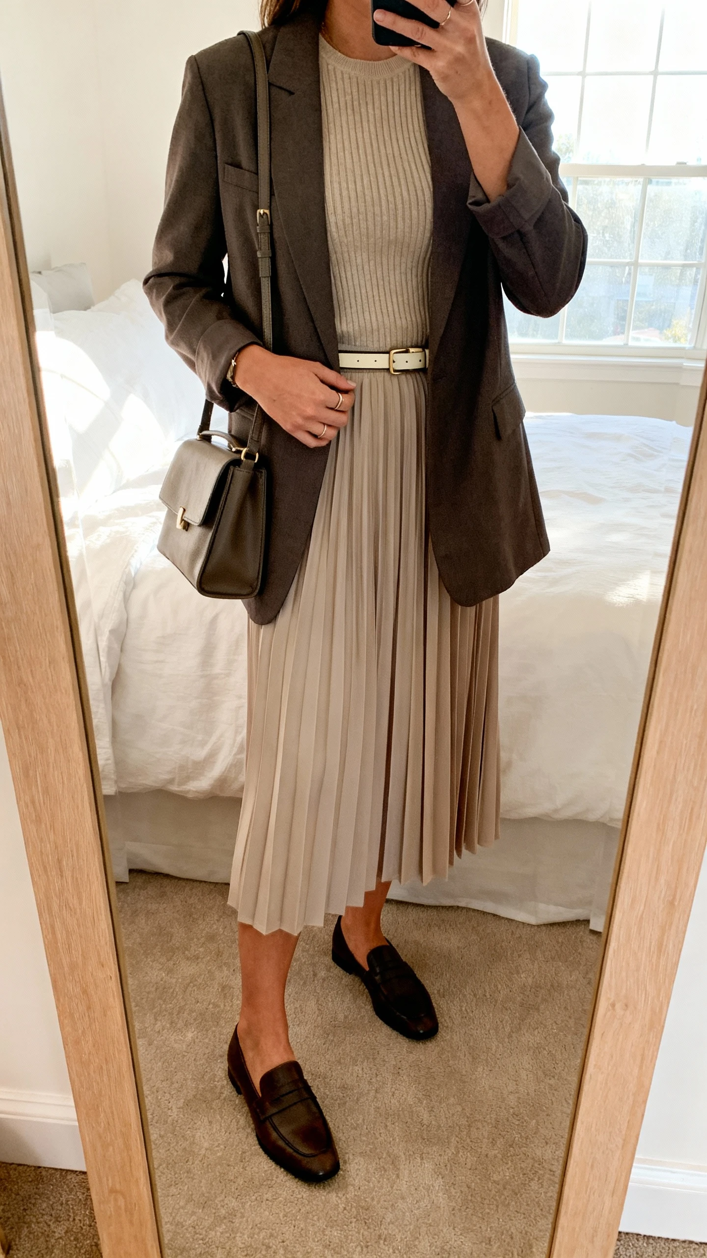 iPhone mirror selfie of a woman wearing a pleated midi skirt with a fitted knit top and a tailored blazer, loafers, structured shoulder bag, and a thin belt, face not visible, cropped at shoulders, bedroom mirror, bright window light, iPhone photo quality.