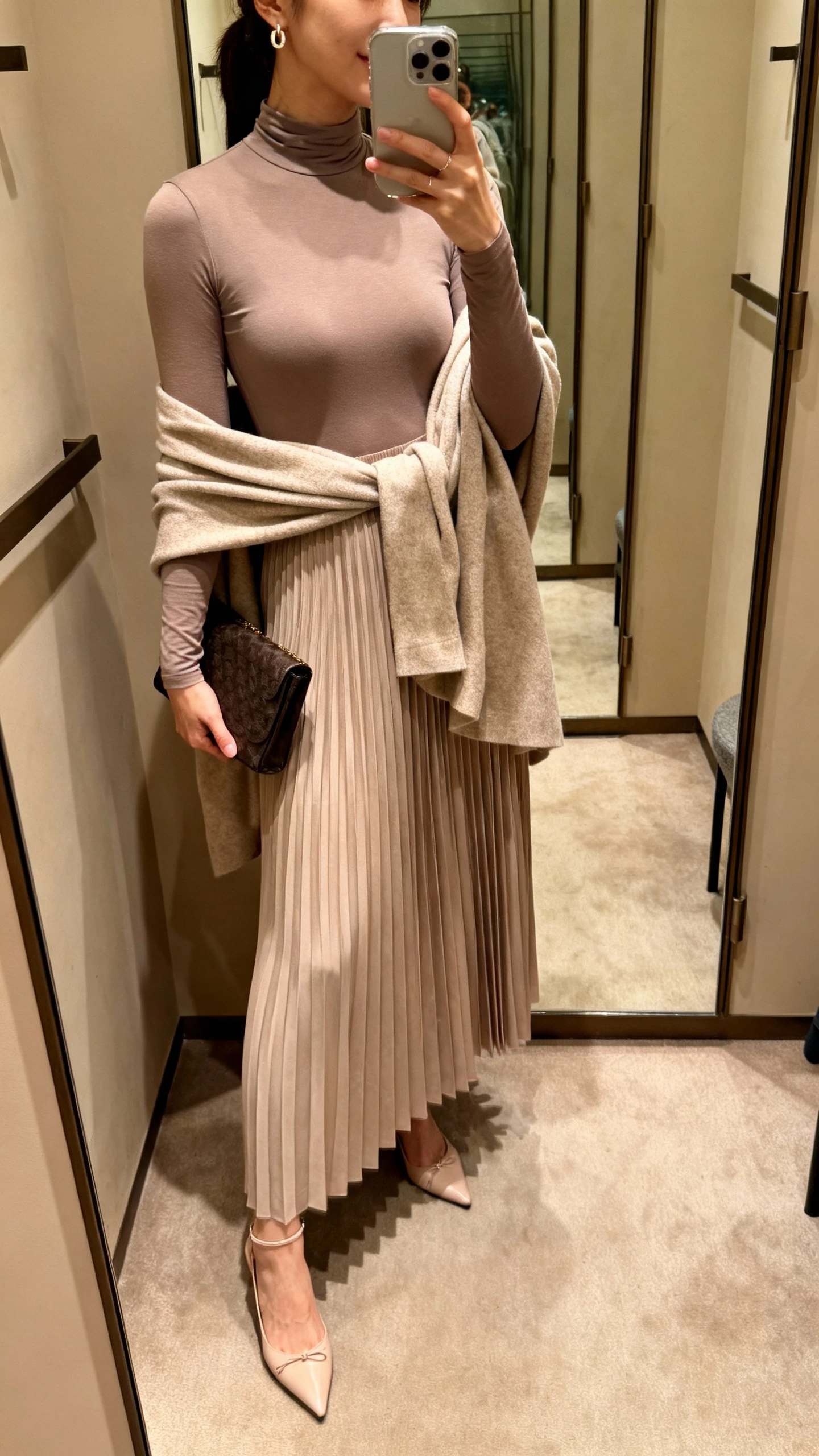 iPhone mirror selfie of a woman wearing a pleated maxi skirt with a modest long-sleeve bodysuit and a draped wrap, delicate heels, clutch bag, and subtle earrings, face not visible, side angle, dressing-room mirror, warm indoor lighting, iPhone photo quality.
