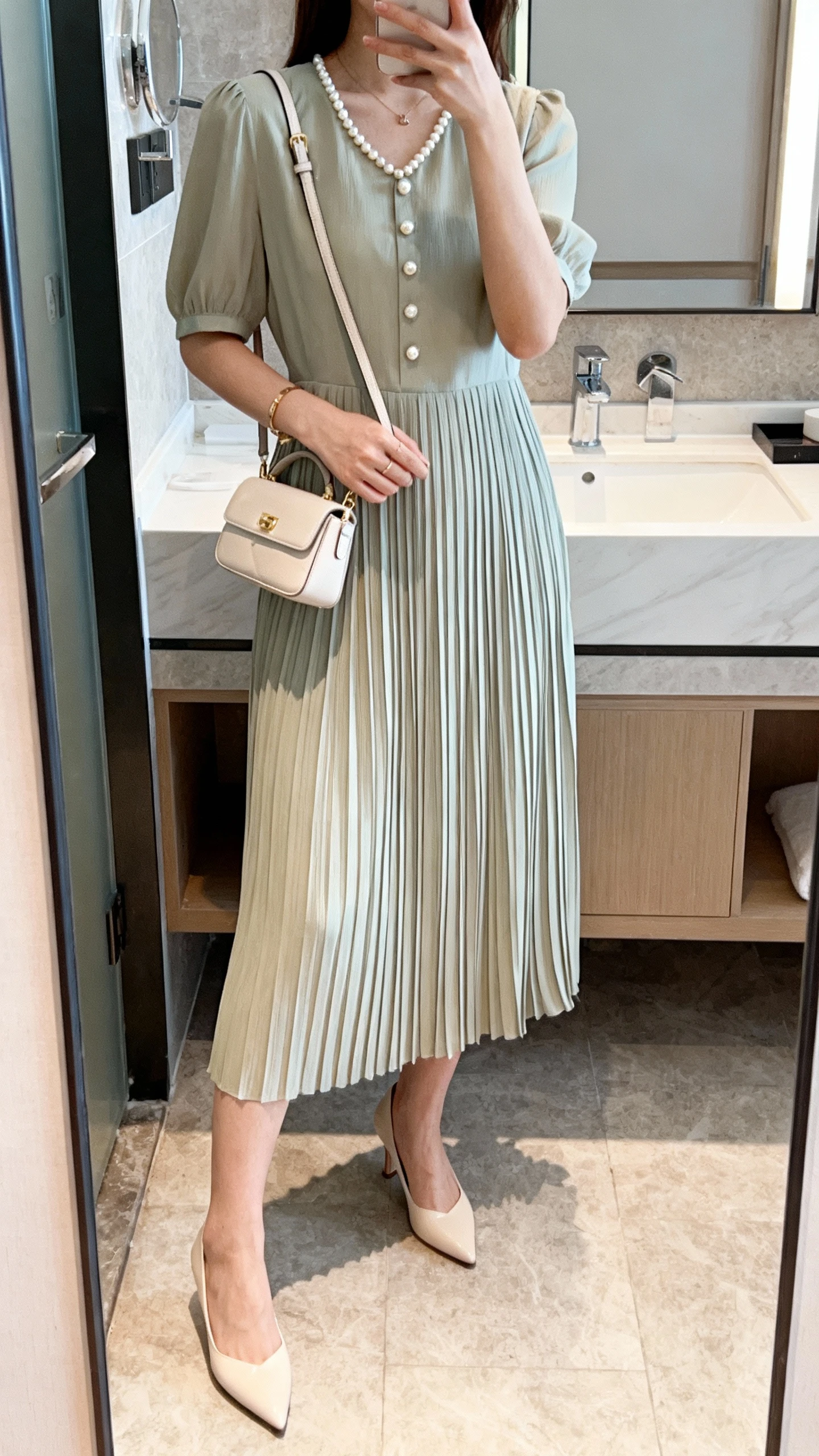 iPhone mirror selfie of a woman wearing a pleated midi dress with pearl button or necklace details, kitten heels, and a petite shoulder bag, face not visible, bathroom mirror, soft indoor lighting, iPhone photo quality.