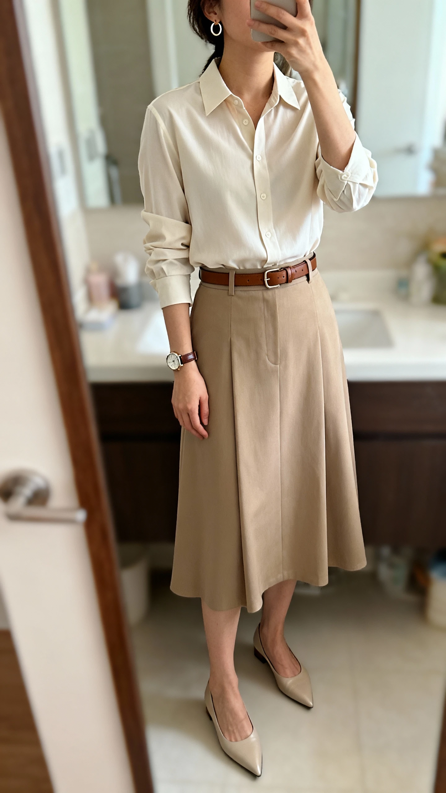 iPhone mirror selfie of a woman wearing a polished blouse tucked into a midi skirt set, slim leather belt, small hoop earrings, classic watch, and pointed-toe flats, face not visible, bathroom full-length mirror, morning indoor lighting, iPhone photo quality.
