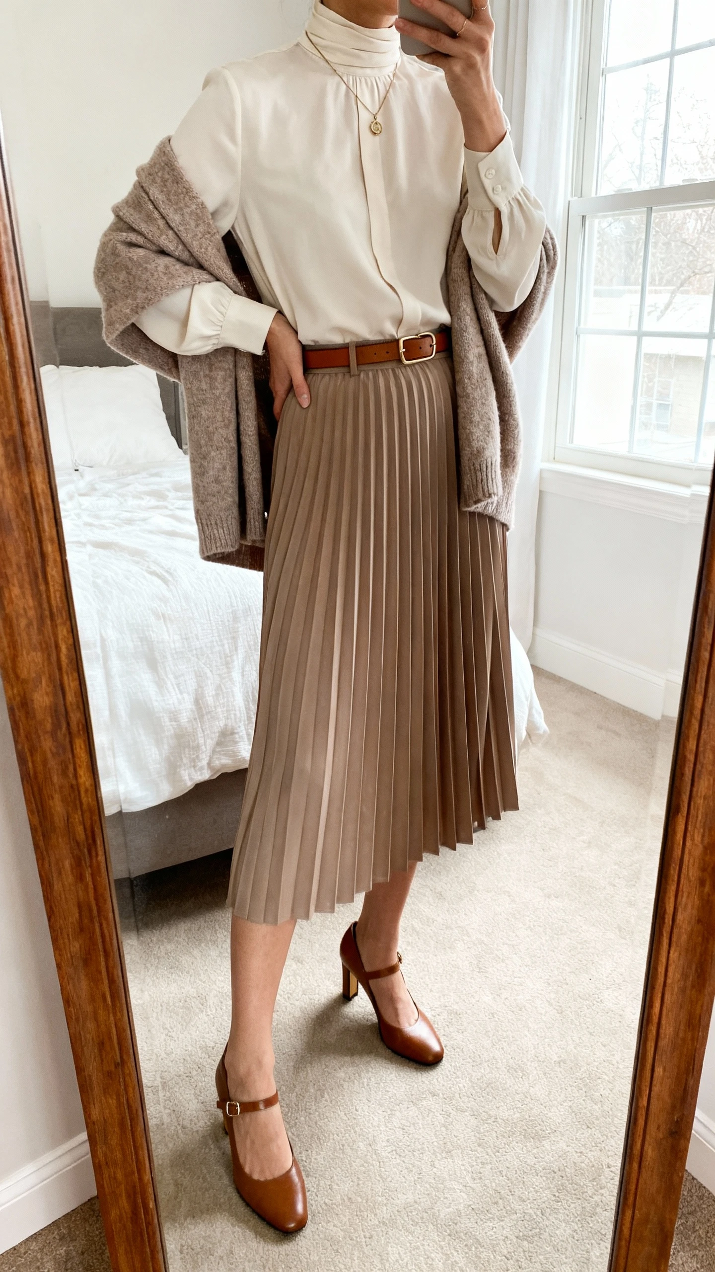 iPhone mirror selfie of a woman wearing a polished high-neck blouse tucked into a pleated midi skirt with a cozy cashmere cardigan draped over shoulders, heeled Mary Janes, delicate pendant, and a slim leather belt, face not visible, cropped at neck, bedroom mirror, natural window light, iPhone photo quality.