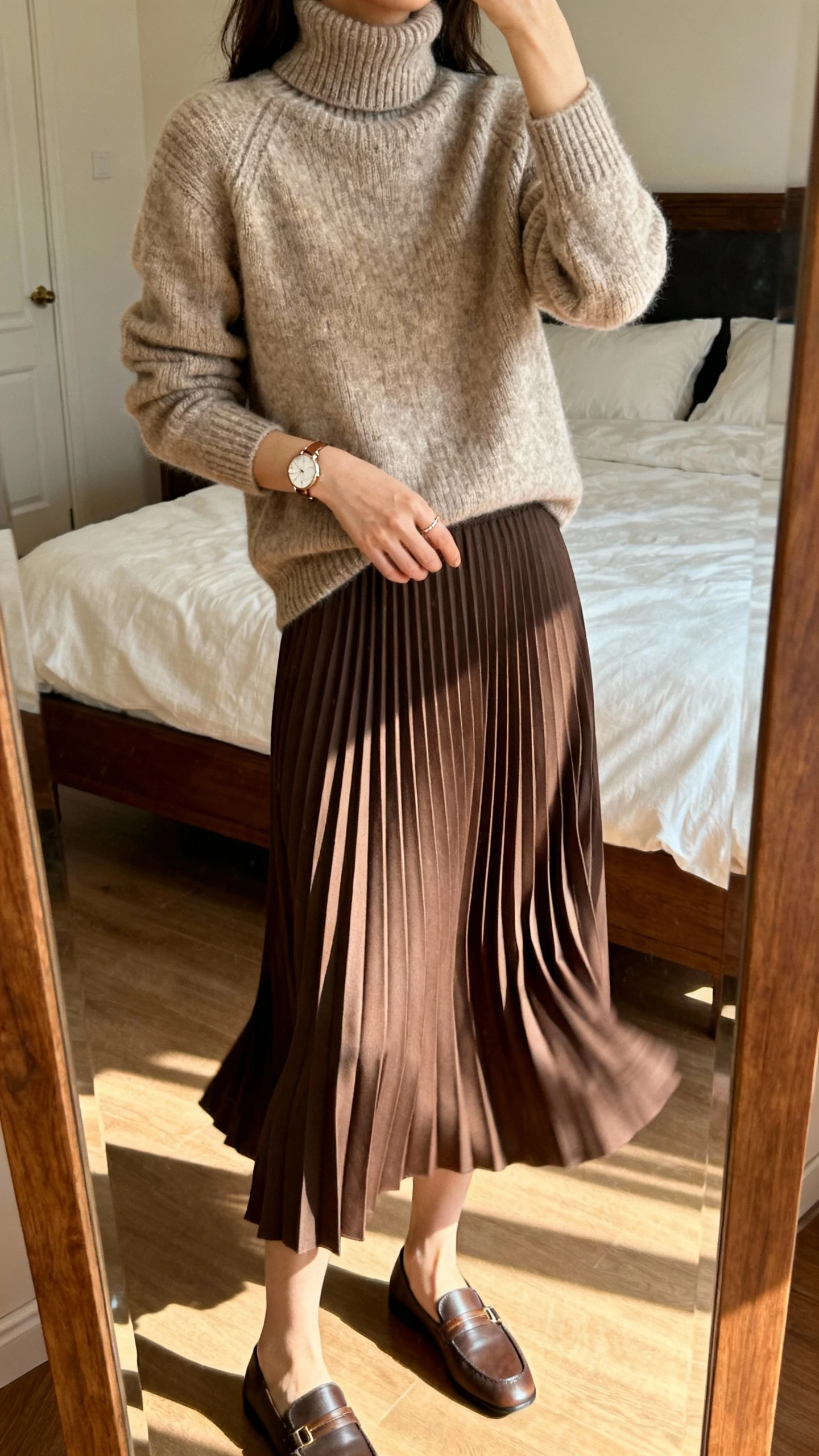 iPhone mirror selfie of a woman wearing a polished knit turtleneck with a swaying pleated skirt, with loafers and a delicate watch, face not visible, cropped at shoulders, bedroom mirror, warm indoor lighting, iPhone photo quality
